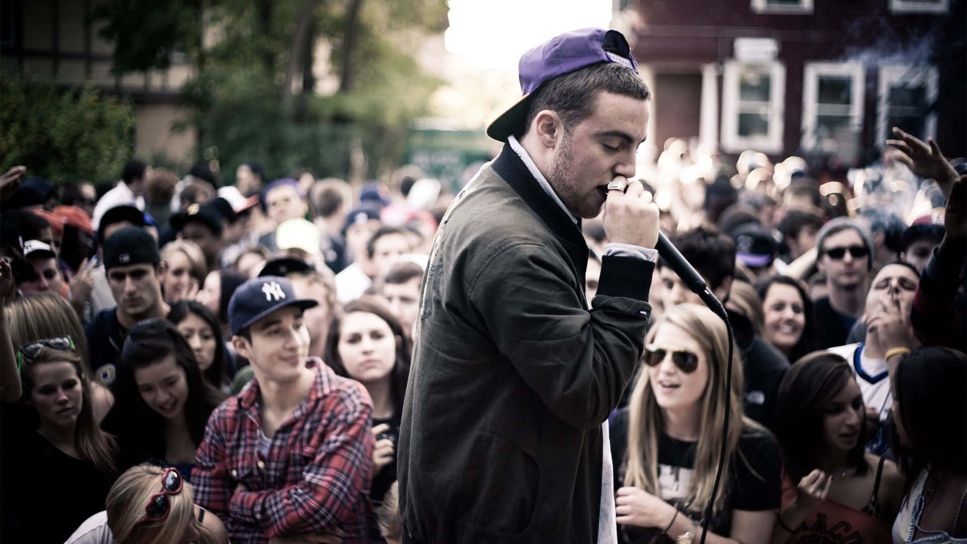 Mac Miller 桌布 壁紙 — 一位戴著紫色帽子的說唱歌手在戶外對著麥克風表演，面前是充滿活力的觀眾。
