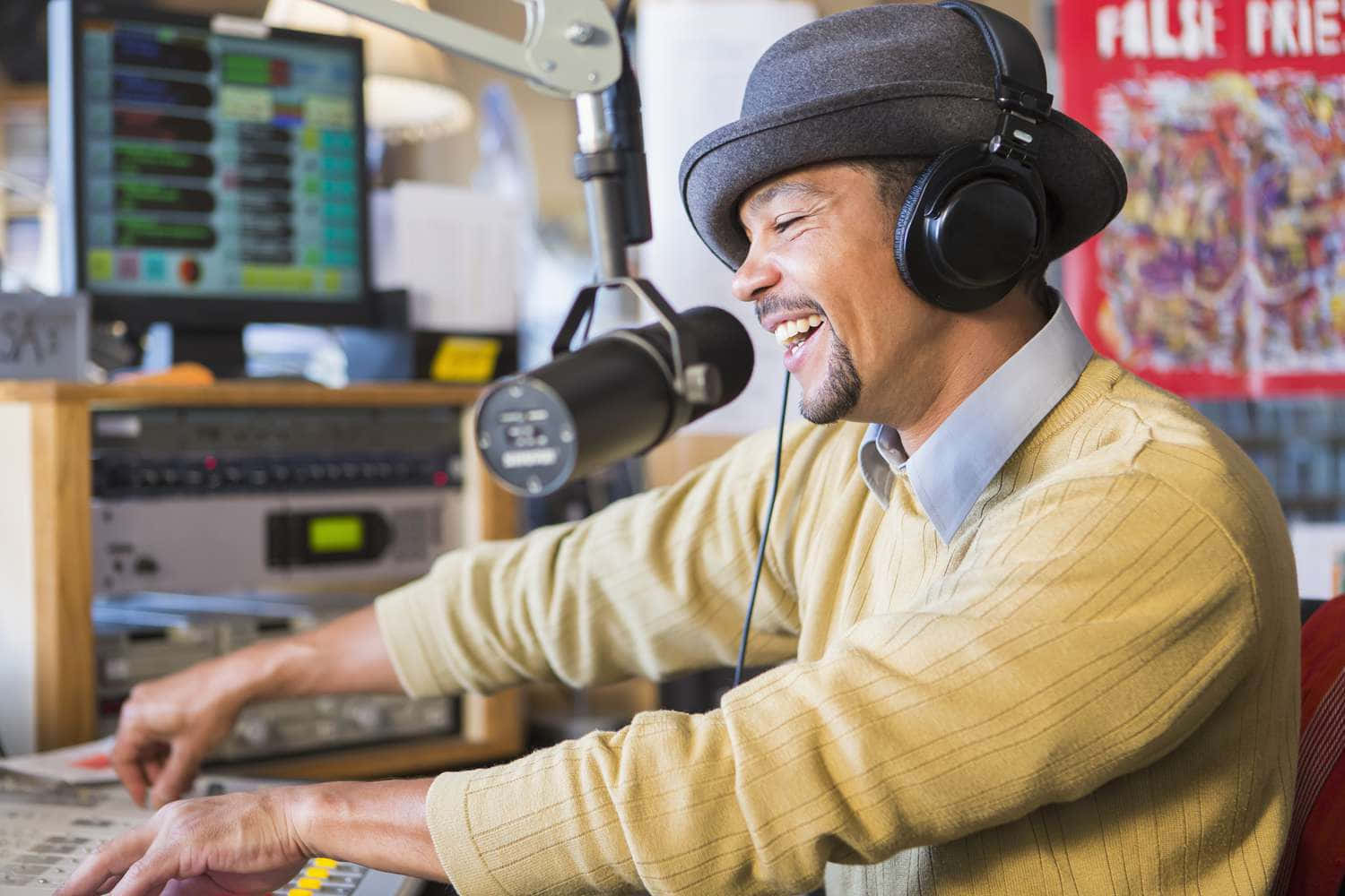 DJ 桌布 壁紙 — 一位戴著紳士帽和耳機的電台主持人微笑著大笑，在錄音室裡廣播，身後有音響設備和彩色海報。