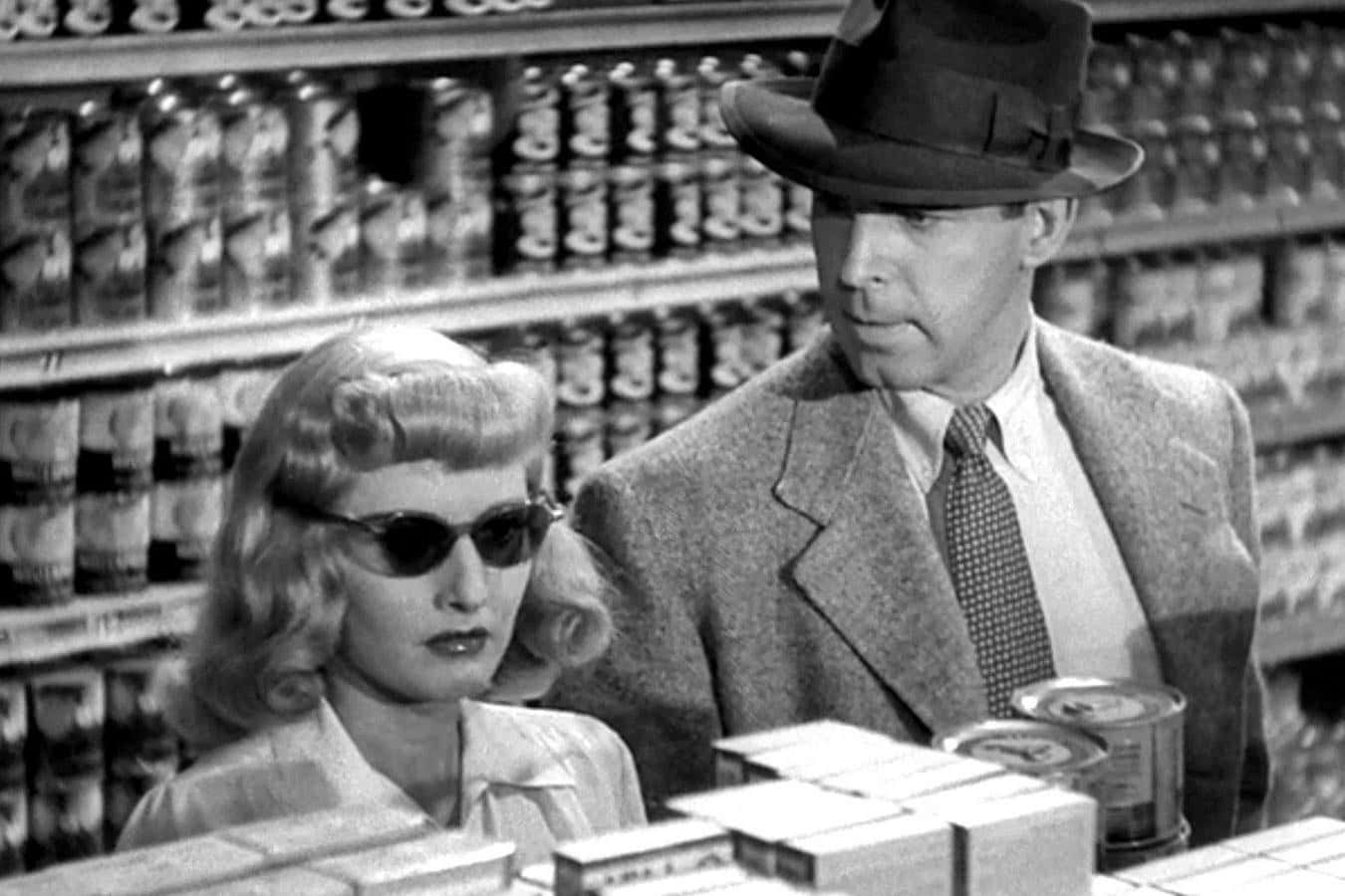 Film Noir 桌布 壁紙 — 一位戴著紳士帽的男人和一位戴著太陽眼鏡的女人站在一個充滿罐頭食品的復古雜貨店走道中。