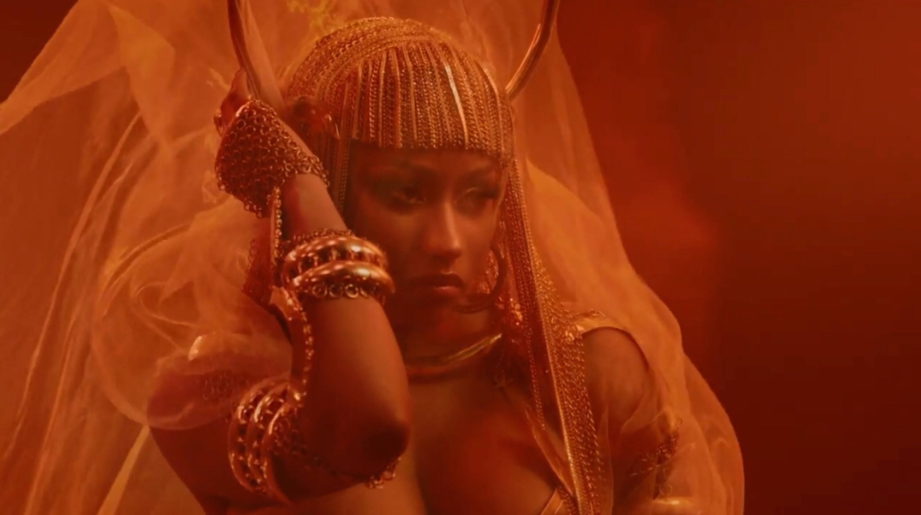 Nicki Minaj Ganja Burn MV 壁紙 — 一位戴著華麗金色頭飾和珠寶的女性，在朦朧的橙色背景下，戴著面紗擺姿勢。