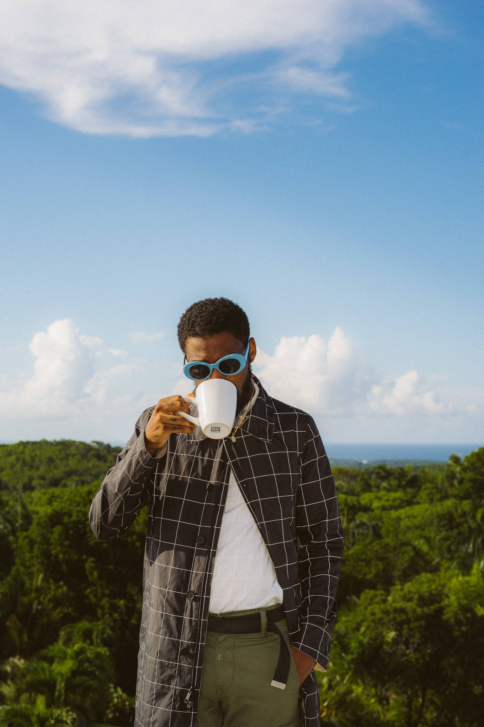 Brent Faiyaz 桌布 壁紙 — 一位戴著藍色太陽眼鏡的時尚男士，在青翠的山丘上俯瞰海洋，在明亮的藍天下啜飲咖啡。