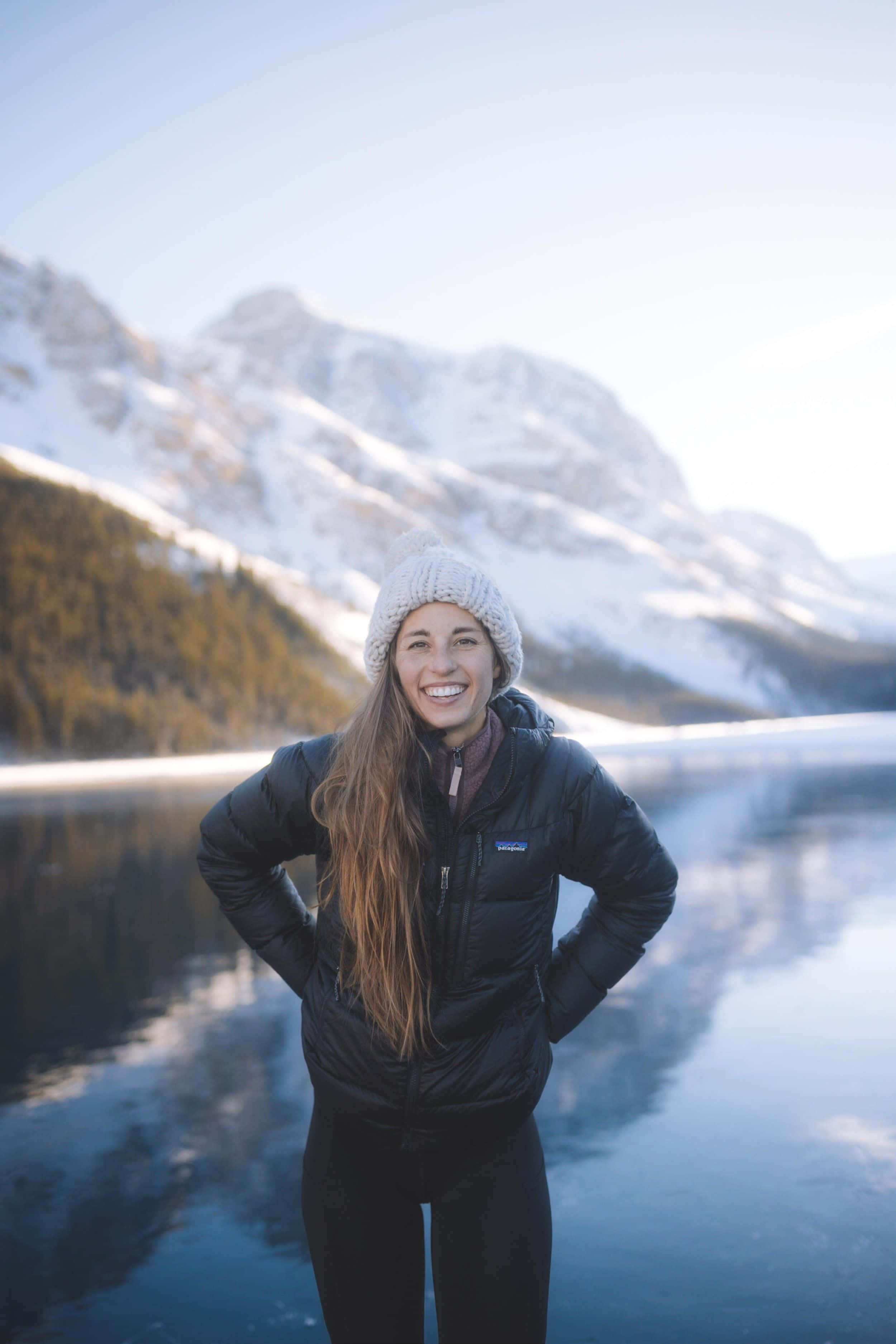 Winter Hiking 桌布 壁紙 — 一位戴著針織帽的微笑女士自信地站在結冰的湖邊，平靜的水面倒映著積雪覆蓋的山脈。