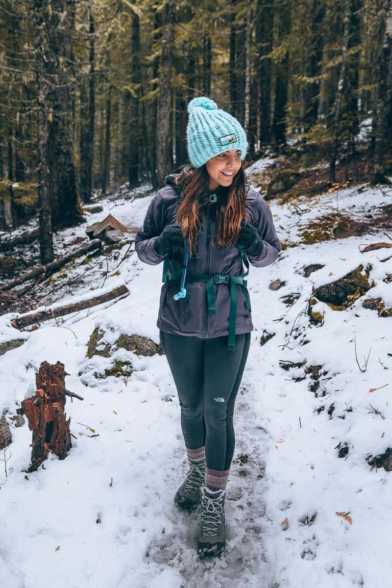 Winter Hiking 桌布 壁紙 — 一位戴著青綠色針織帽的微笑女性，在白雪覆蓋的森林小徑上徒步，周圍是高聳的松樹。