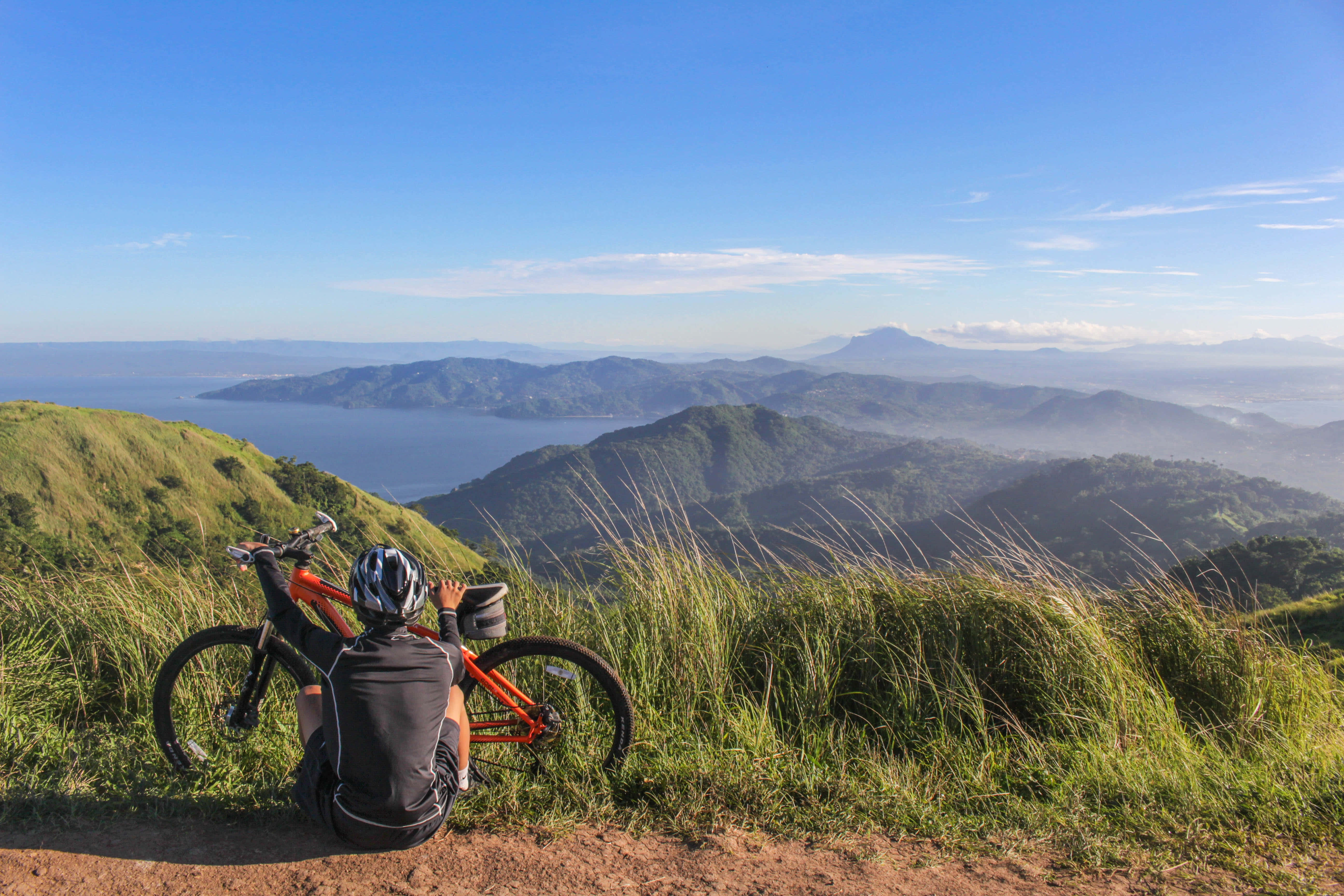 Outdoor Activities 桌布 壁紙 — 一位戴著頭盔的單車手在長滿草的山頂上，在他們的橙色單車旁邊休息，俯瞰著廣闊的海洋和層層疊疊的山脈，天空清澈湛藍。