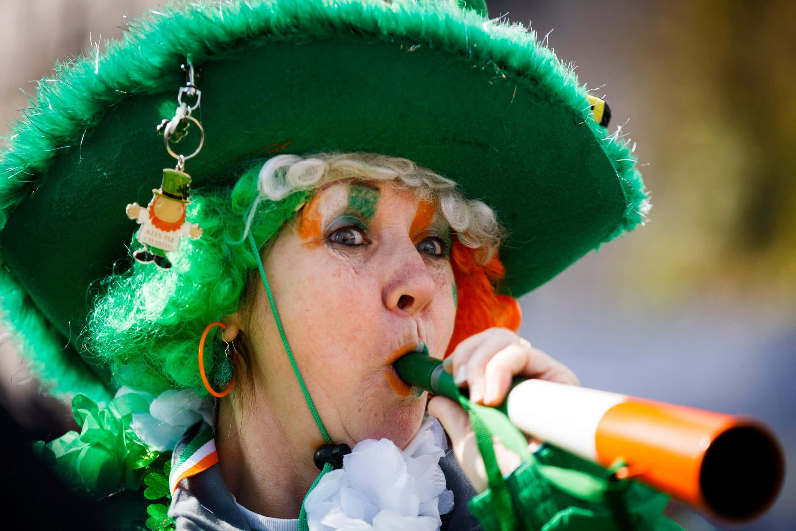 St Patrick's Day 桌布 壁紙 — 一位戴著鮮綠色帽子和臉部彩繪的女性，在聖派翠克節慶祝活動中演奏節日愛爾蘭號角。