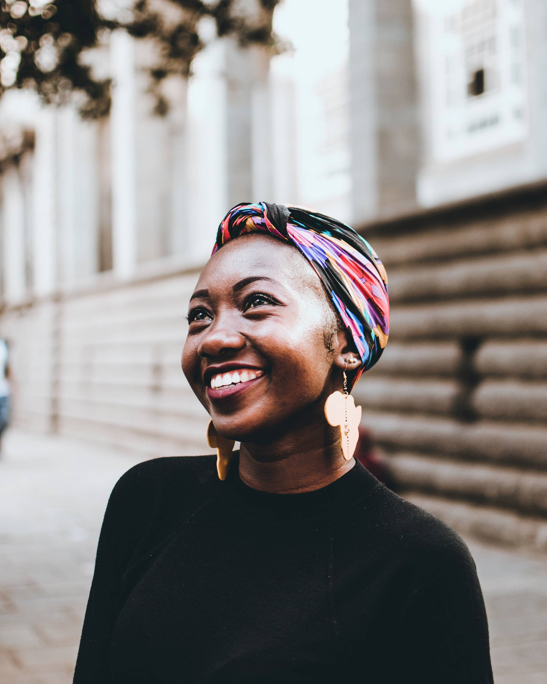 empowered black woman with vibrant head wrap 壁紙 — 一位戴著鮮豔頭巾的快樂女性，在陽光明媚的城市街道上行走時，露出燦爛的笑容。