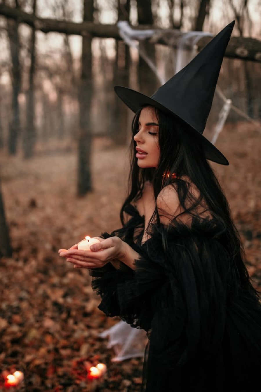 Witch Costumes 桌布 壁紙 — 一位戴著黑色帽子的女巫在迷霧繚繞的秋日森林中手持一支點燃的蠟燭，地上散落著落葉和發光的蠟燭。