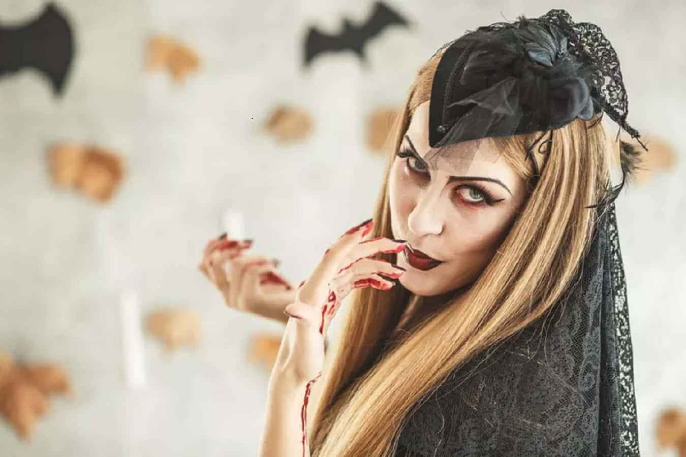 Vampire Costumes 桌布 壁紙 — 一位戴著黑色蕾絲面紗和帽子、手上沾滿鮮血的女性，在裝飾有蝙蝠和南瓜的牆前擺姿勢。