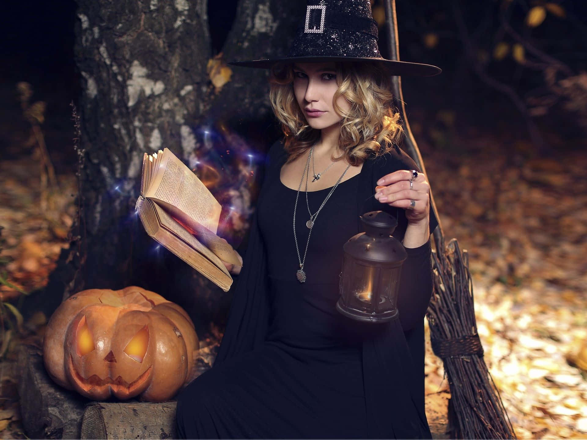 Witches Broom 桌布 壁紙 — 一位戴黑帽穿黑裙的女巫坐在暮色中的森林裡，從一本打開的書中施展魔法，旁邊有一個發光的傑克南瓜燈。