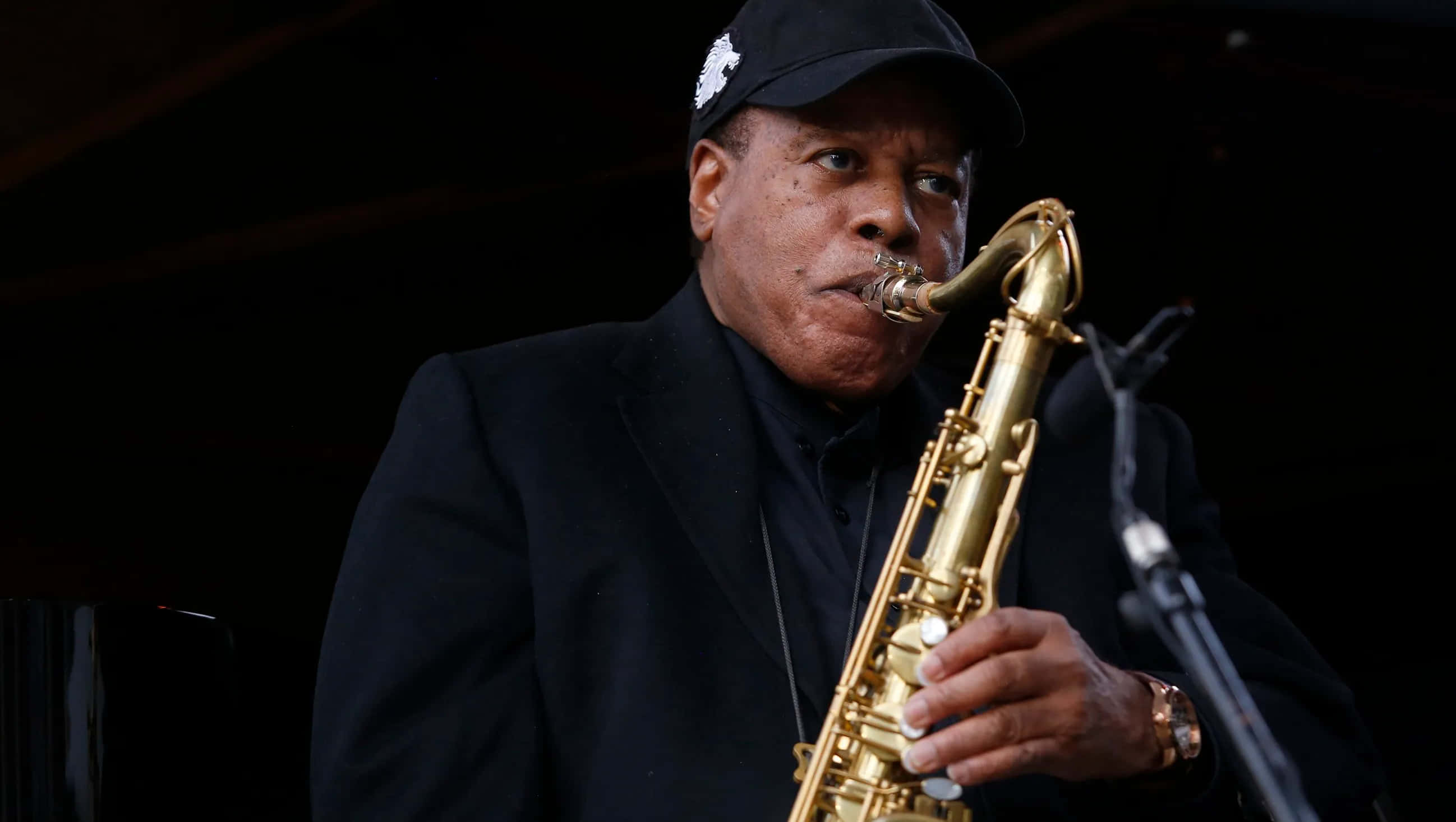 Wayne Shorter 桌布 壁紙 — 一位戴黑色帽子和西裝的爵士音樂家在舞台燈光下熱情地吹奏金色色士風。