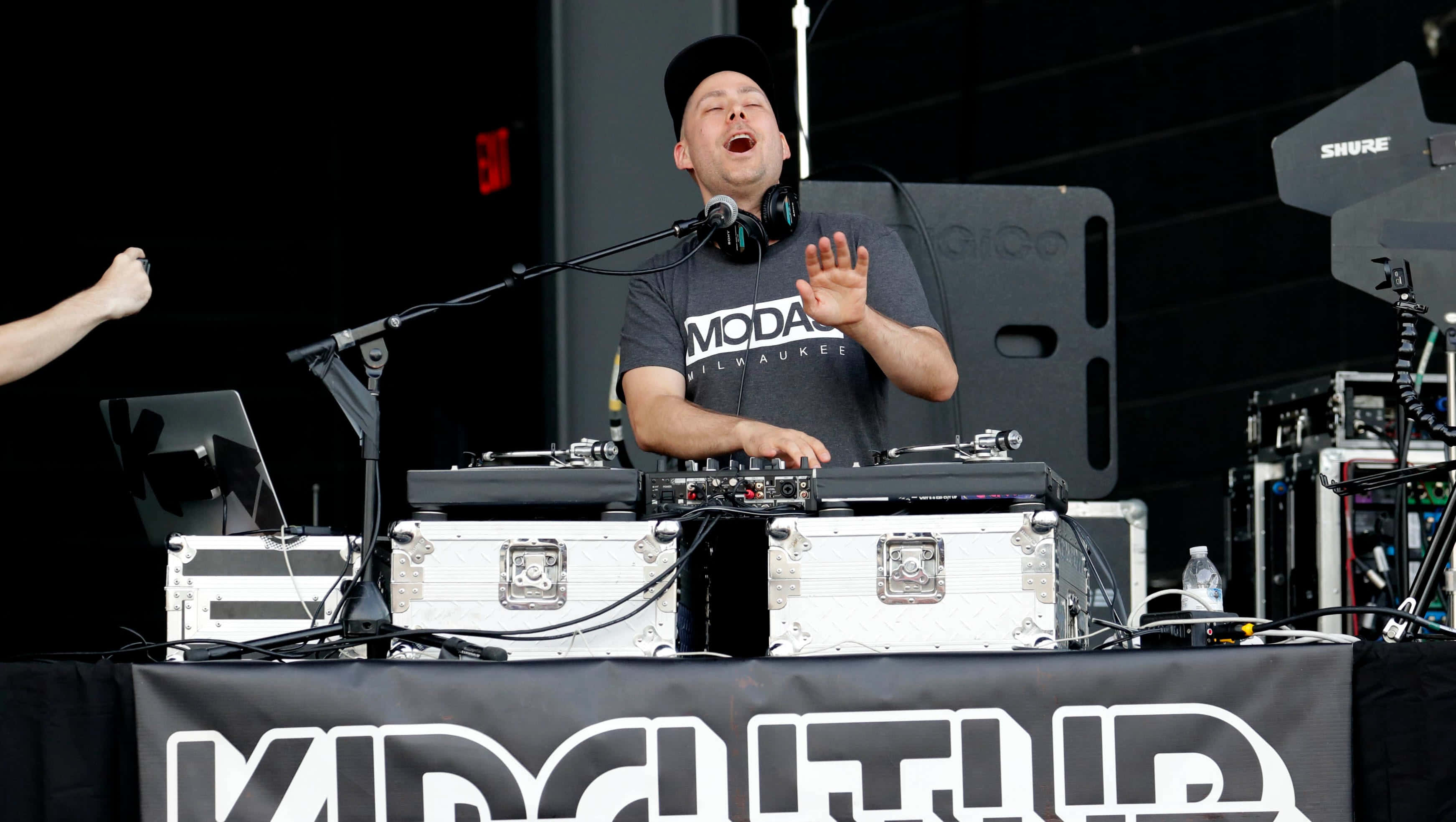 DJ 桌布 壁紙 — 一位戴黑色鴨舌帽、穿灰色襯衫的DJ在音樂節上熱情表演，雙手高舉，耳機掛在脖子上。