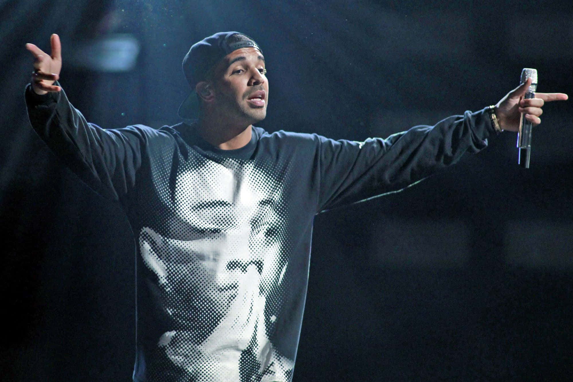 Drake 桌布 壁紙 — 一位戴黑色鴨舌帽、身穿圖案衛衣的男表演者，在黑暗的舞台上張開雙臂握著麥克風，充滿激情地演唱。