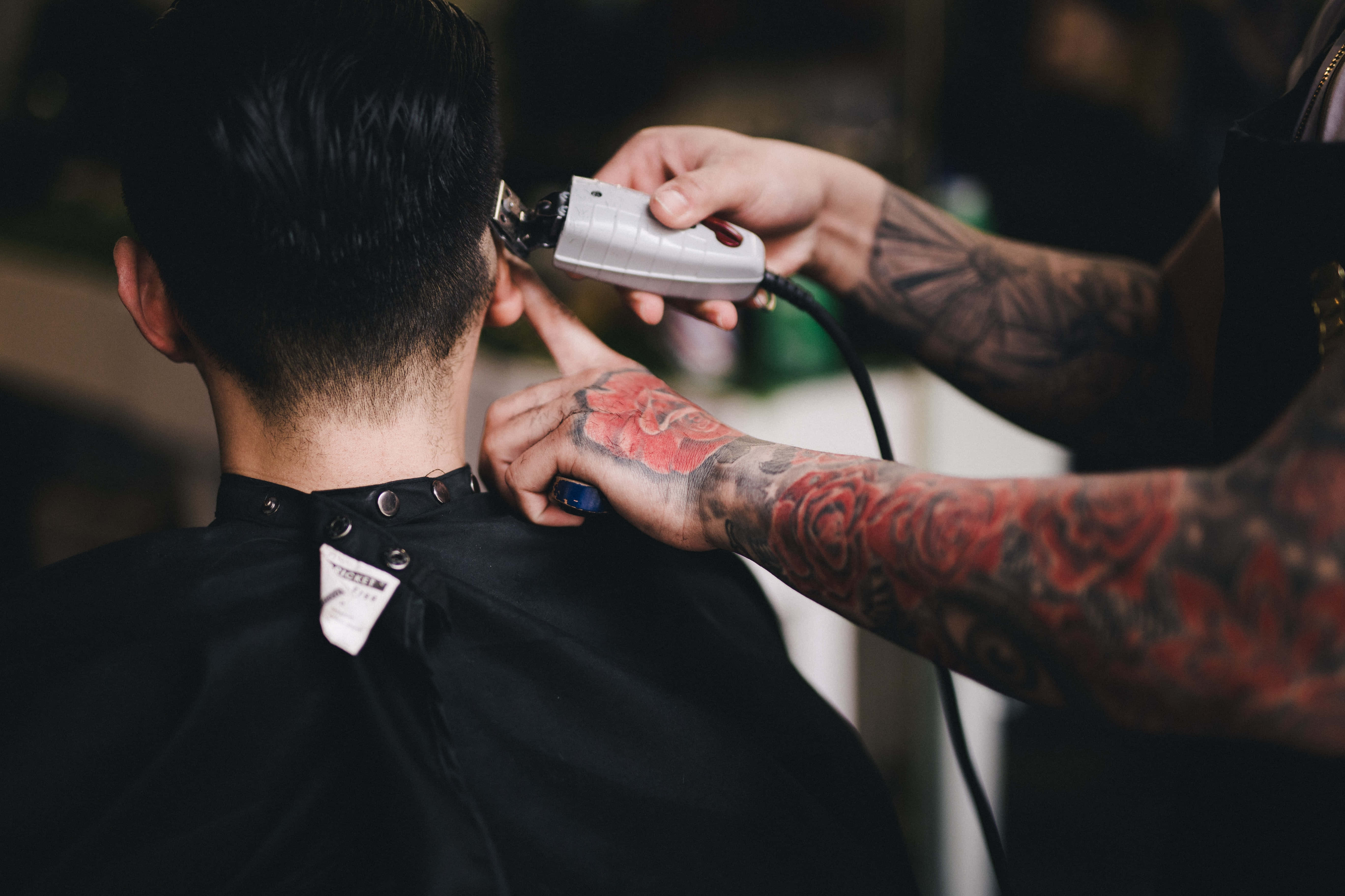 Barber 桌布 壁紙 — 一位手臂有紋身的理髮師在髮廊用電動推剪修剪顧客的短黑髮。