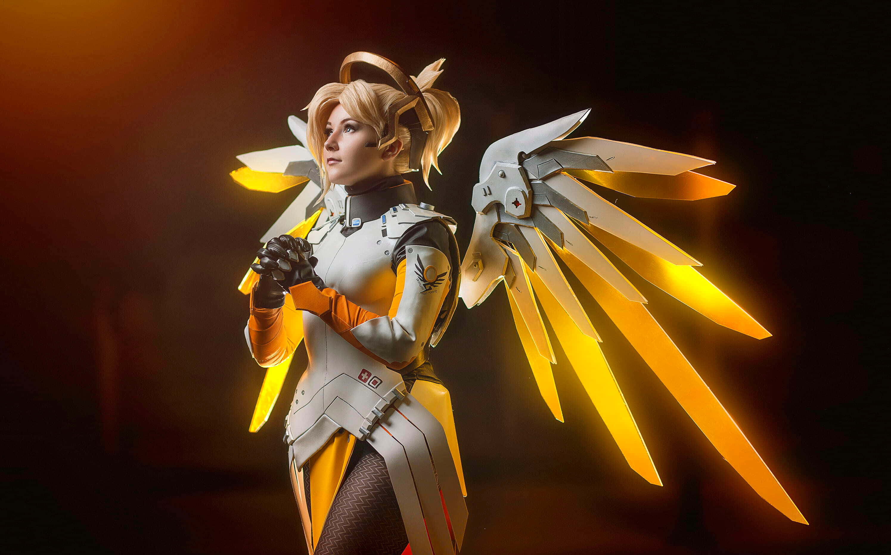 Gaming Cosplay 桌布 壁紙 — 一位扮演《鬥陣特攻》慈悲的Cosplayer，在昏暗溫暖的燈光背景下，展開閃耀的金色翅膀。