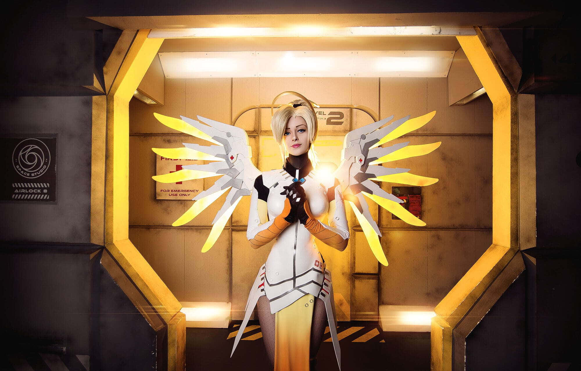 Gaming Cosplay 桌布 壁紙 — 一位扮演《鬥陣特攻》英雄慈悲的cosplayer站在發光的黃色六邊形門口，手持發光的白色和黃色翅膀。