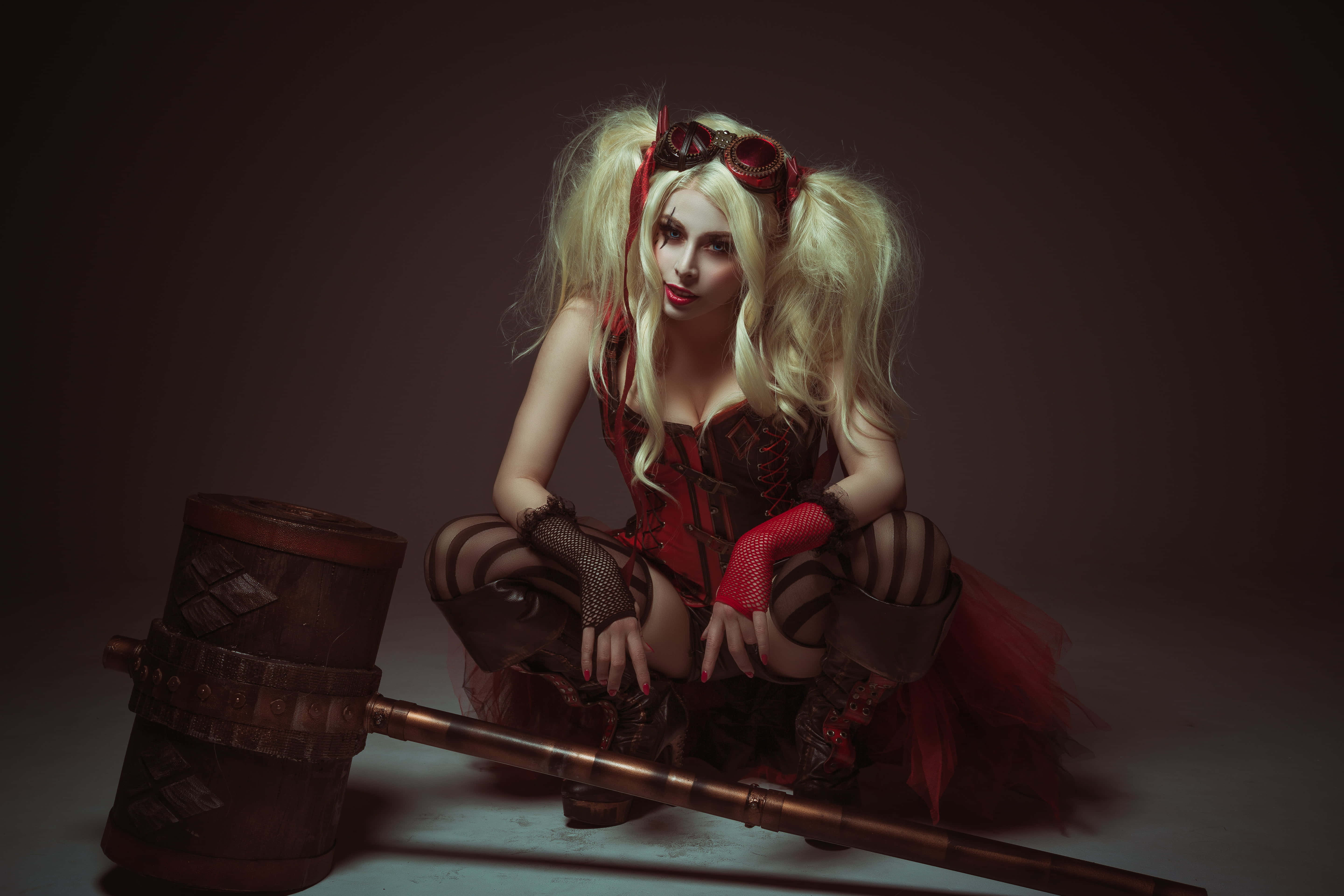 Harley Quinn Hammer 桌布 壁紙 — 一位扮演哈莉·奎茵的角色扮演者蹲在一個黑暗的攝影棚裡，旁邊放著一個大槌，燈光效果戲劇化。