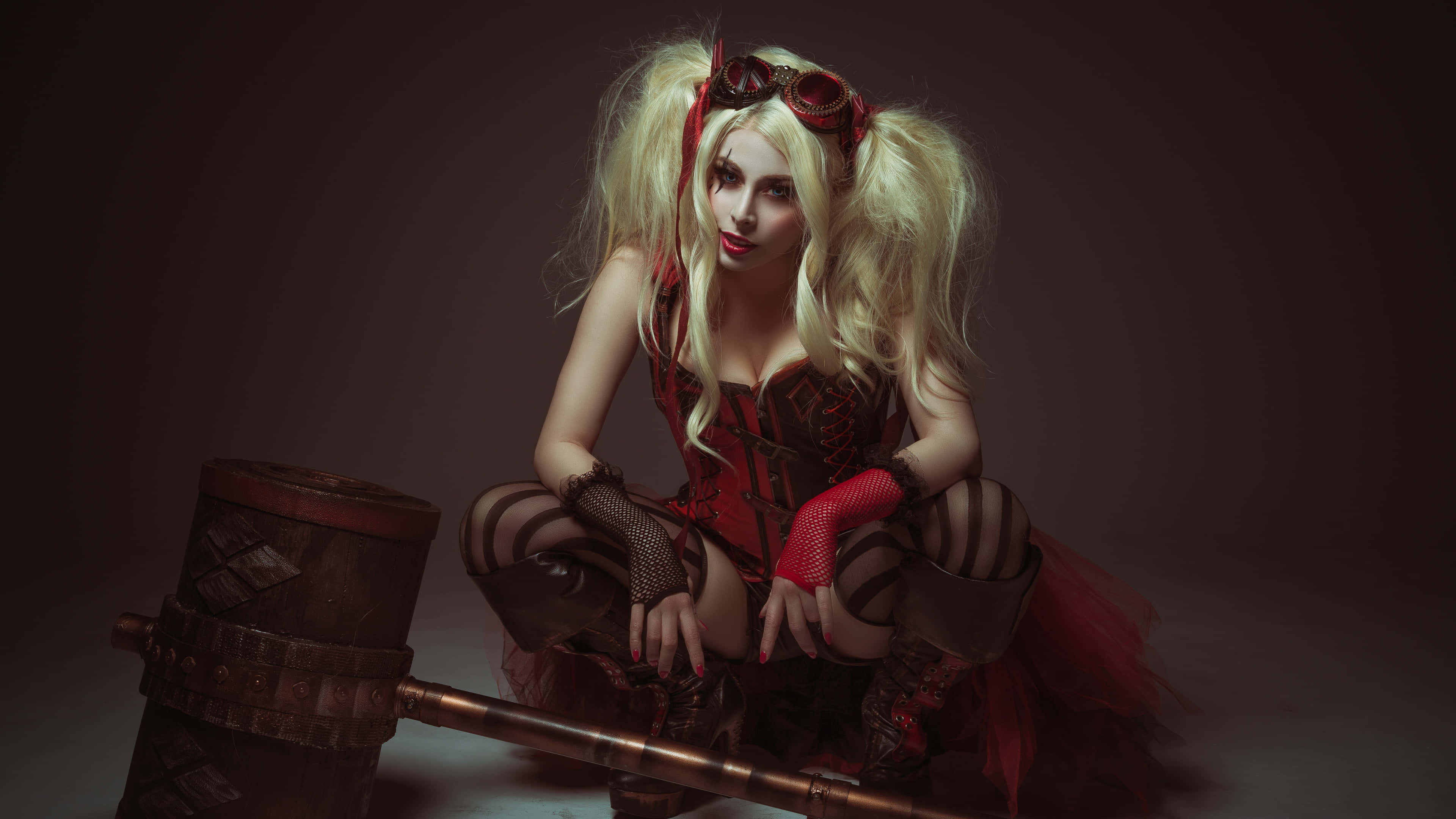 Harley Quinn Cosplay 桌布 壁紙 — 一位扮演小丑女哈莉·奎茵的角色扮演者，在昏暗的攝影棚中，伴隨著戲劇性的紅色燈光，蹲在一個巨大的槌子旁邊。