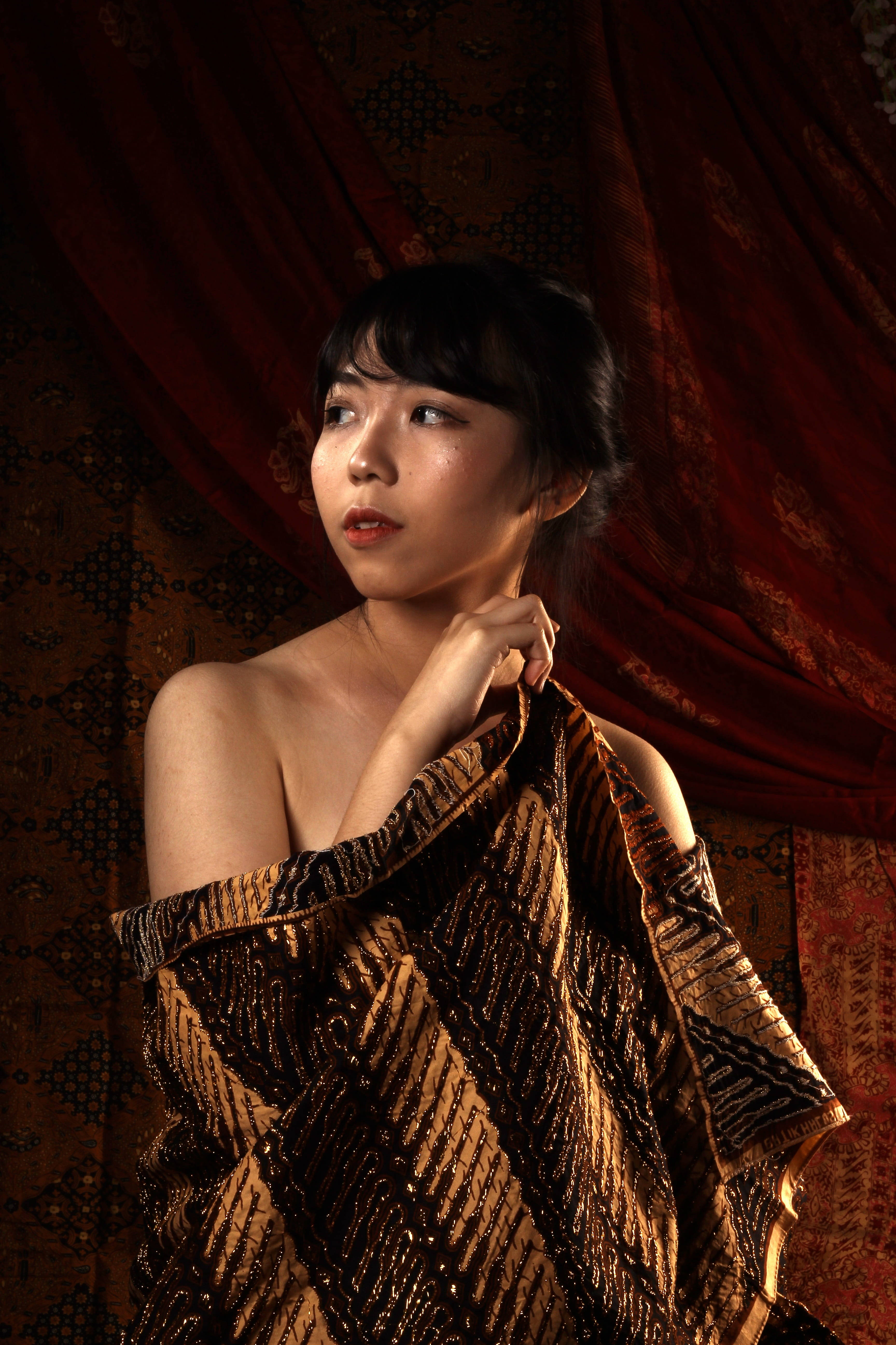 Clothes 桌布 壁紙 — 一位披著金色圖案布料的女性，在溫暖的攝影棚燈光下，仰望著華麗的紅色天鵝絨窗簾。