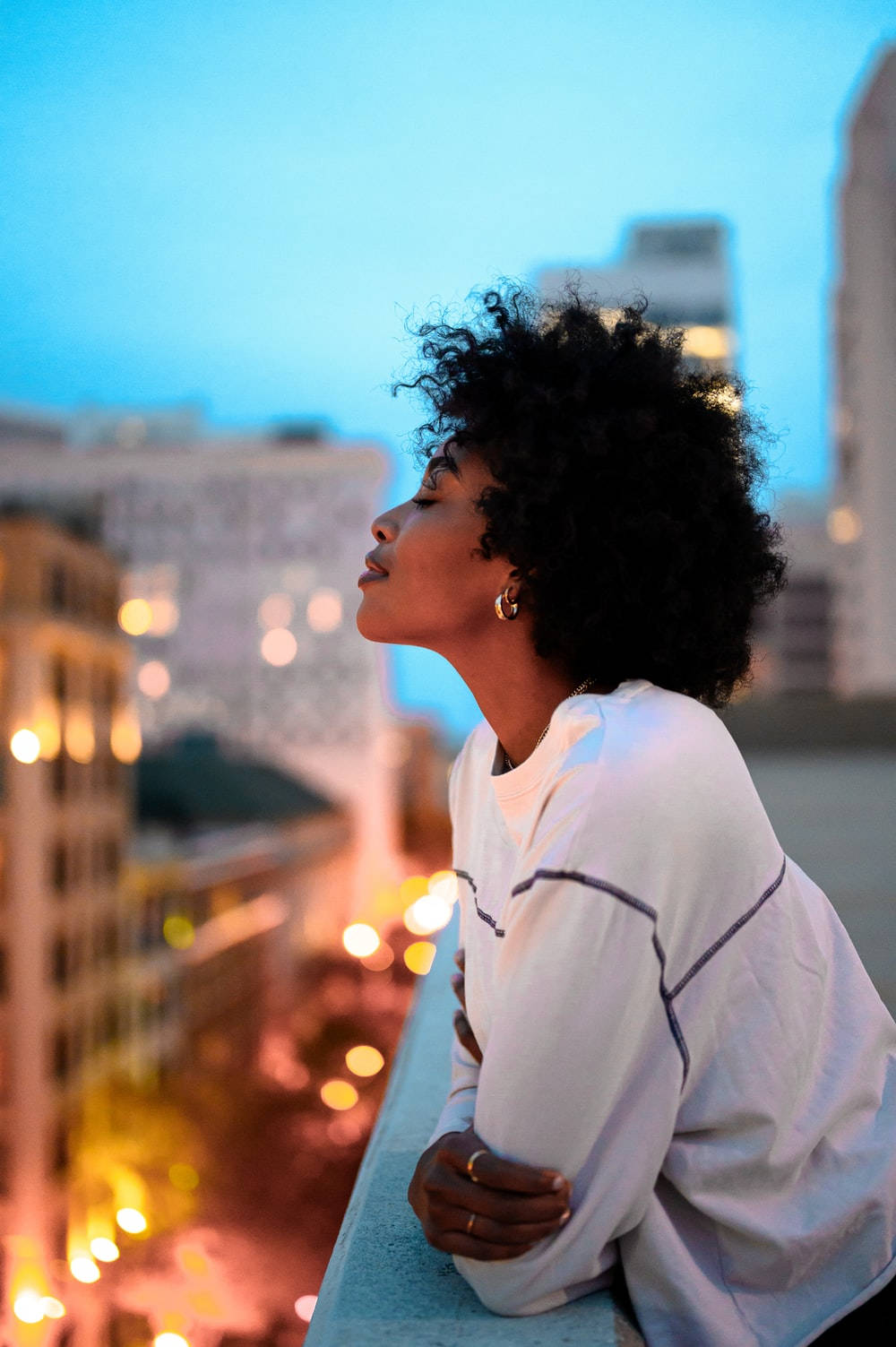 empowered black woman embracing the night city lights 壁紙 — 一位捲髮女性在屋頂上仰望，城市燈光在暮色天空中模糊成溫暖的散景。