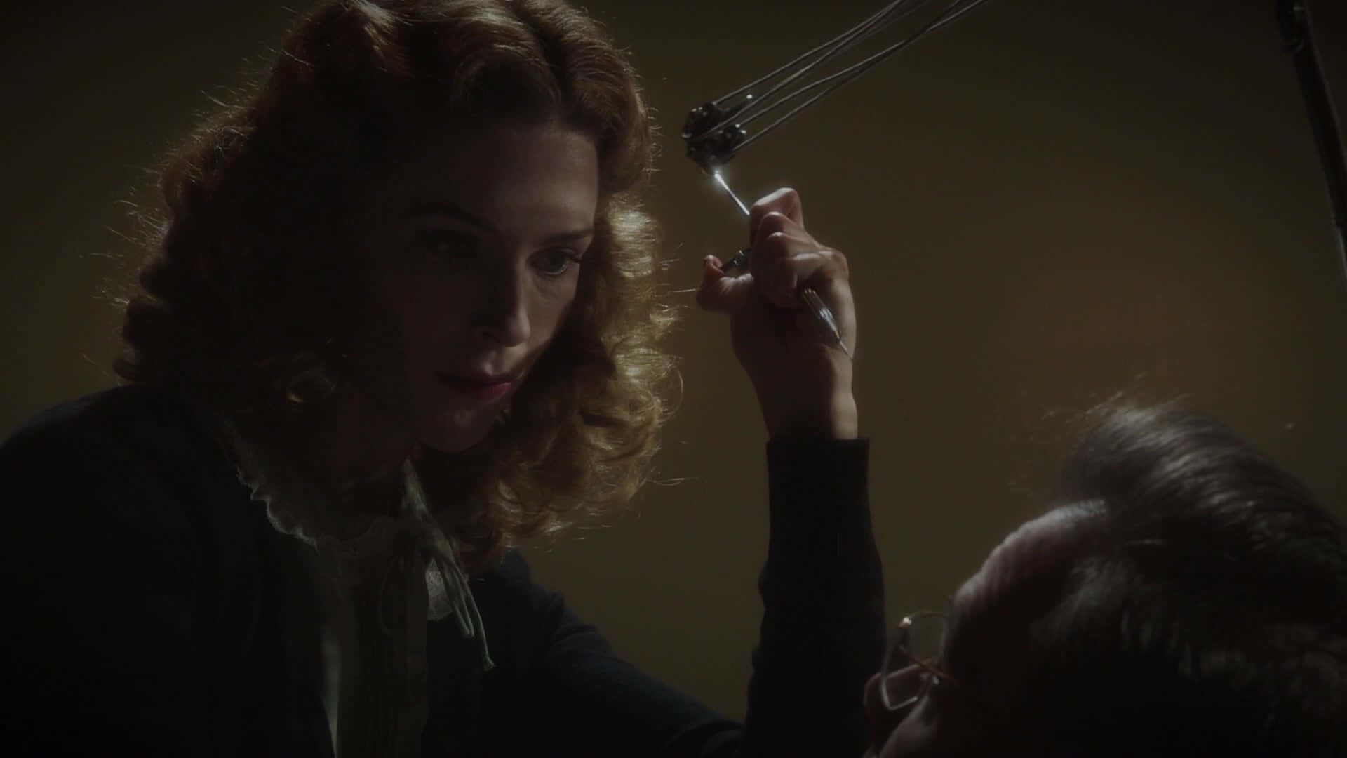 Peggy Carter 桌布 壁紙 — 一位捲髮女性在昏暗的房間裡，將手術工具靠近另一個人的頭部。