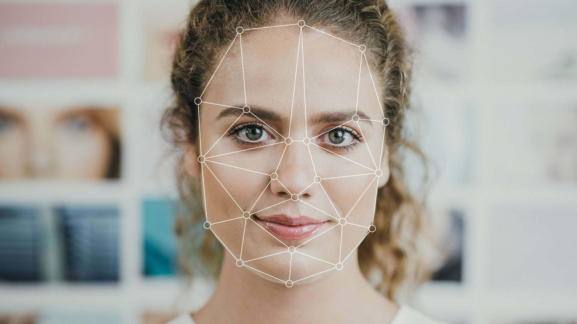 Face Recognition 桌布 壁紙 — 一位捲髮女性微笑，白色幾何面部識別網格疊加在她的臉上，背景是模糊的照片牆。