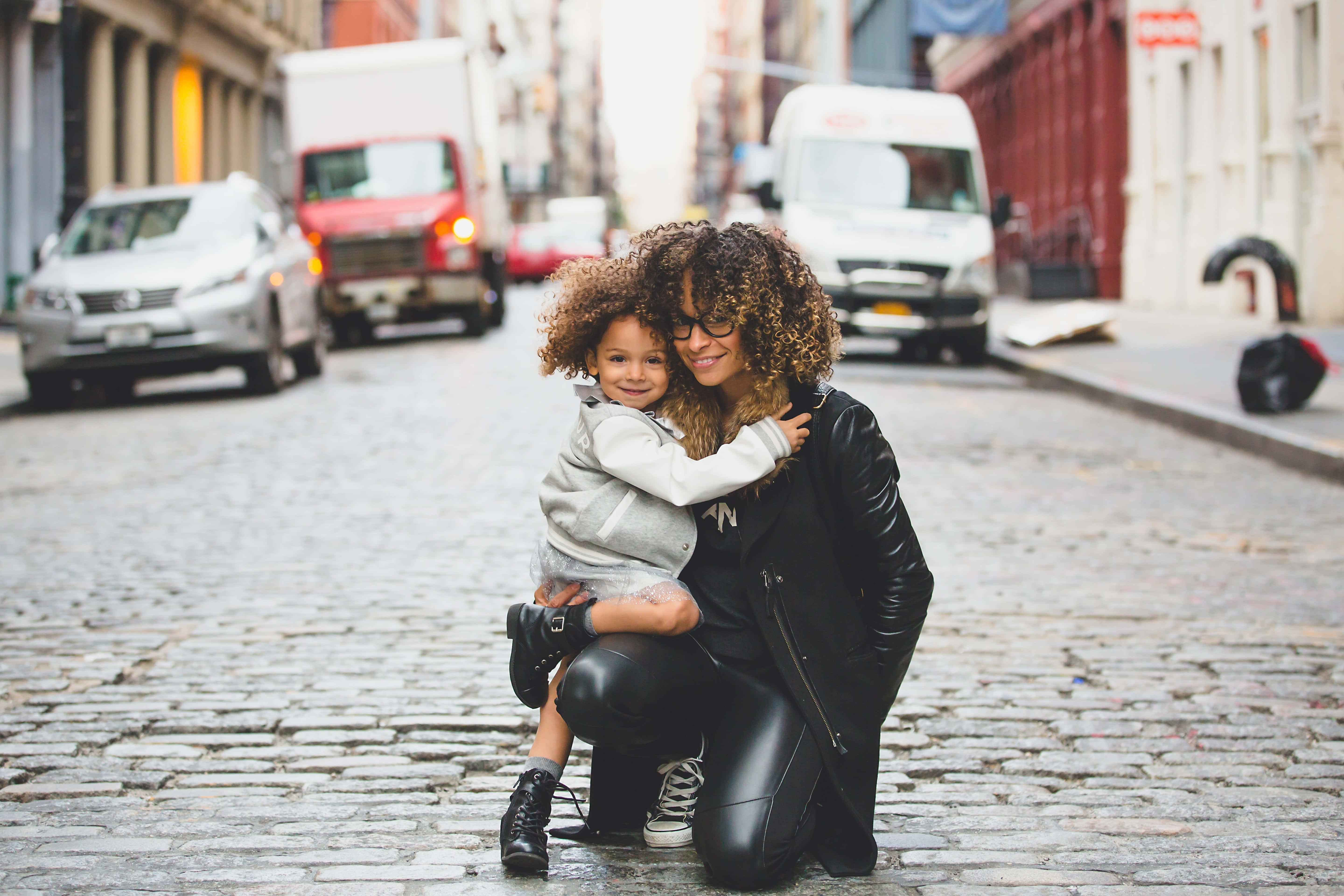 Mom 桌布 壁紙 — 一位捲髮女性跪在鵝卵石街道上，抱著一個穿著灰色連帽衫、微笑的孩子。