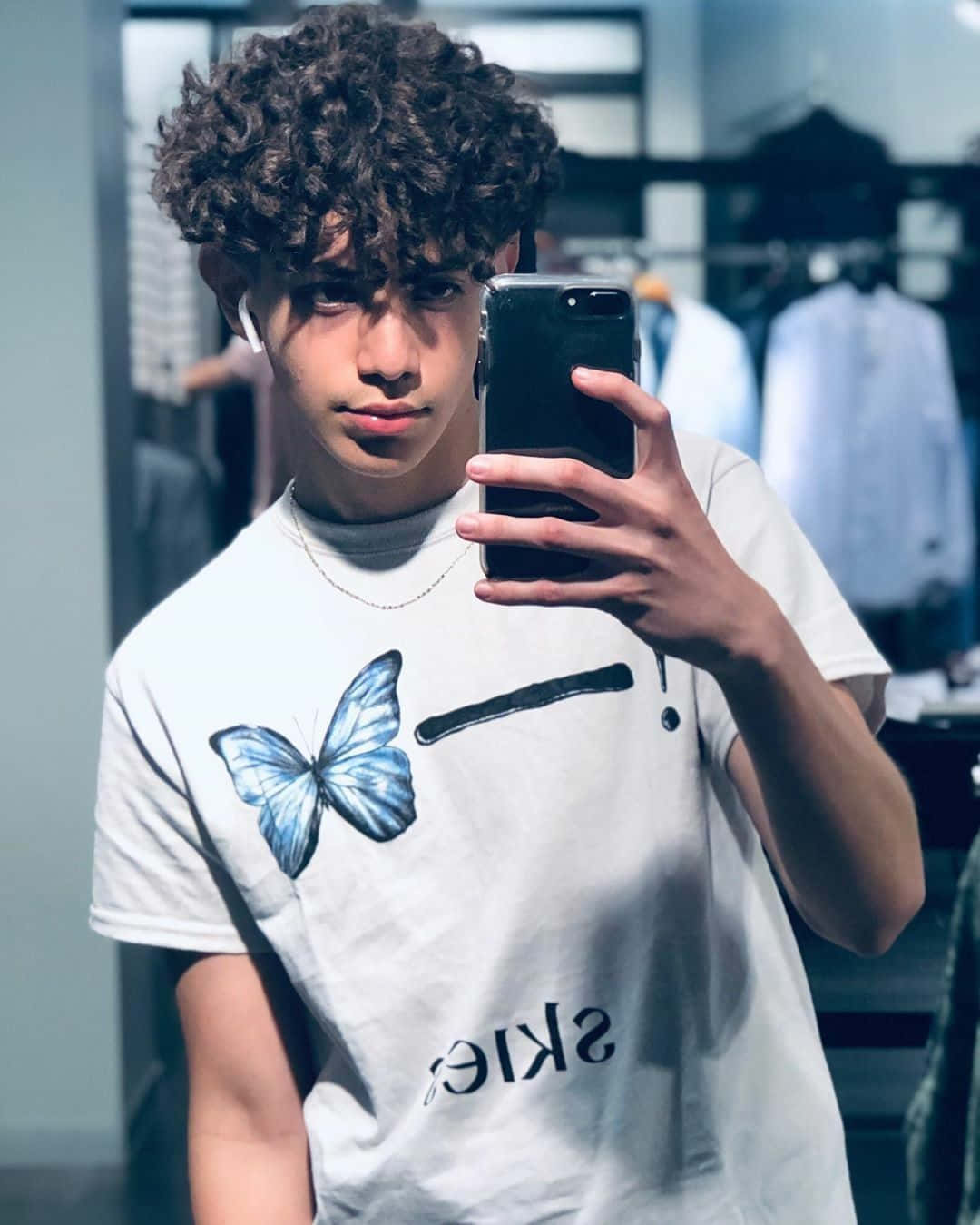 Boy 桌布 壁紙 — 一位捲髮年輕男子在服裝店穿著帶有藍色蝴蝶圖案的白色T恤，對著鏡子自拍。