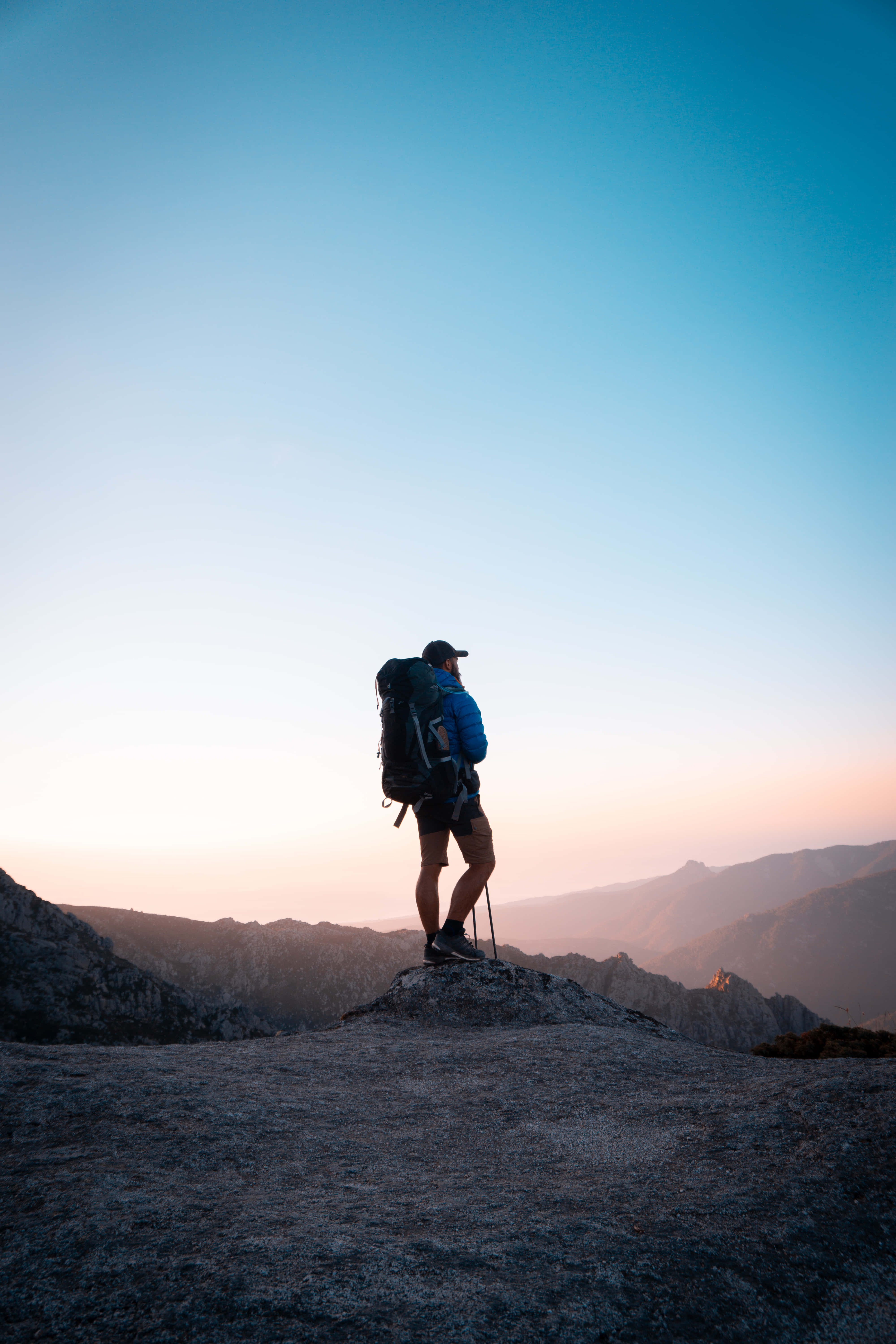 Hiking 桌布 壁紙 — 一位揹著大背包的遠足者站在岩石山頂上，在日出時分俯瞰漸變天空下的遠處山脊。