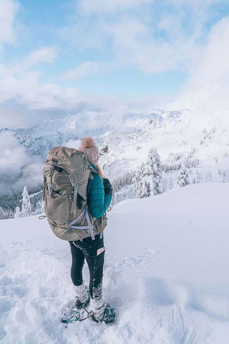 Winter Hiking 桌布 壁紙 — 一位揹著大背包的遠足者站在白雪覆蓋的山頂上，俯瞰著部分多雲天空下廣闊的冬季景觀。