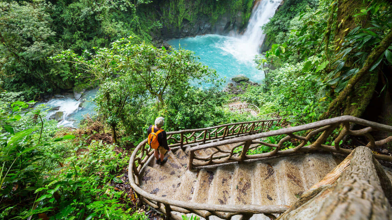Costa Rica 桌布 壁紙 — 一位揹著橙色背包的遠足者，沿著一條蜿蜒穿過茂密綠色叢林的質樸木樓梯，走向一個碧綠色的瀑布。