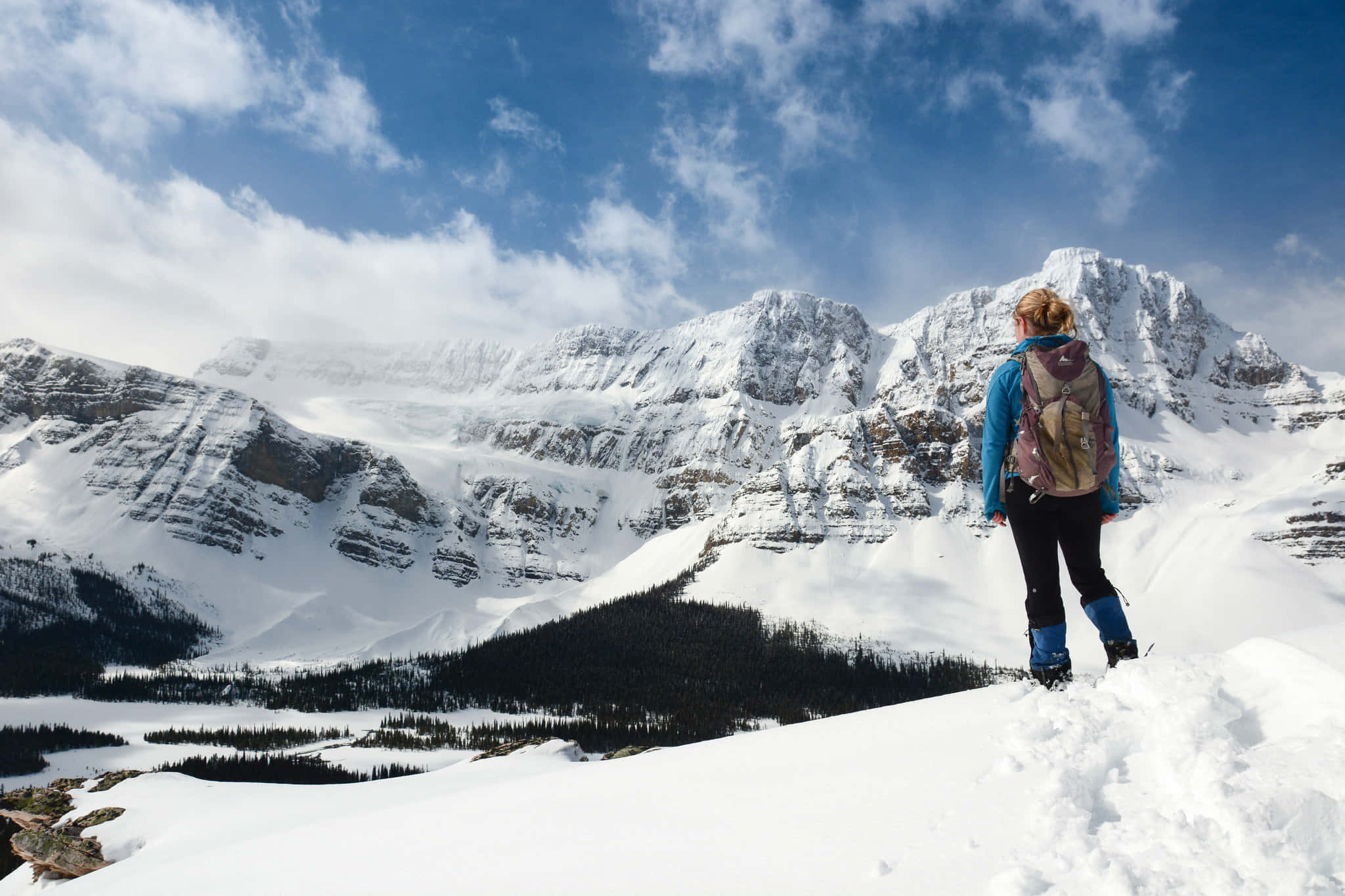 Hiking 桌布 壁紙 — 一位揹著背包的遠足者站在白雪覆蓋的山脊上，俯瞰廣闊的白雪覆蓋的山谷和參差不齊的山峰，天空湛藍，雲朵點綴。