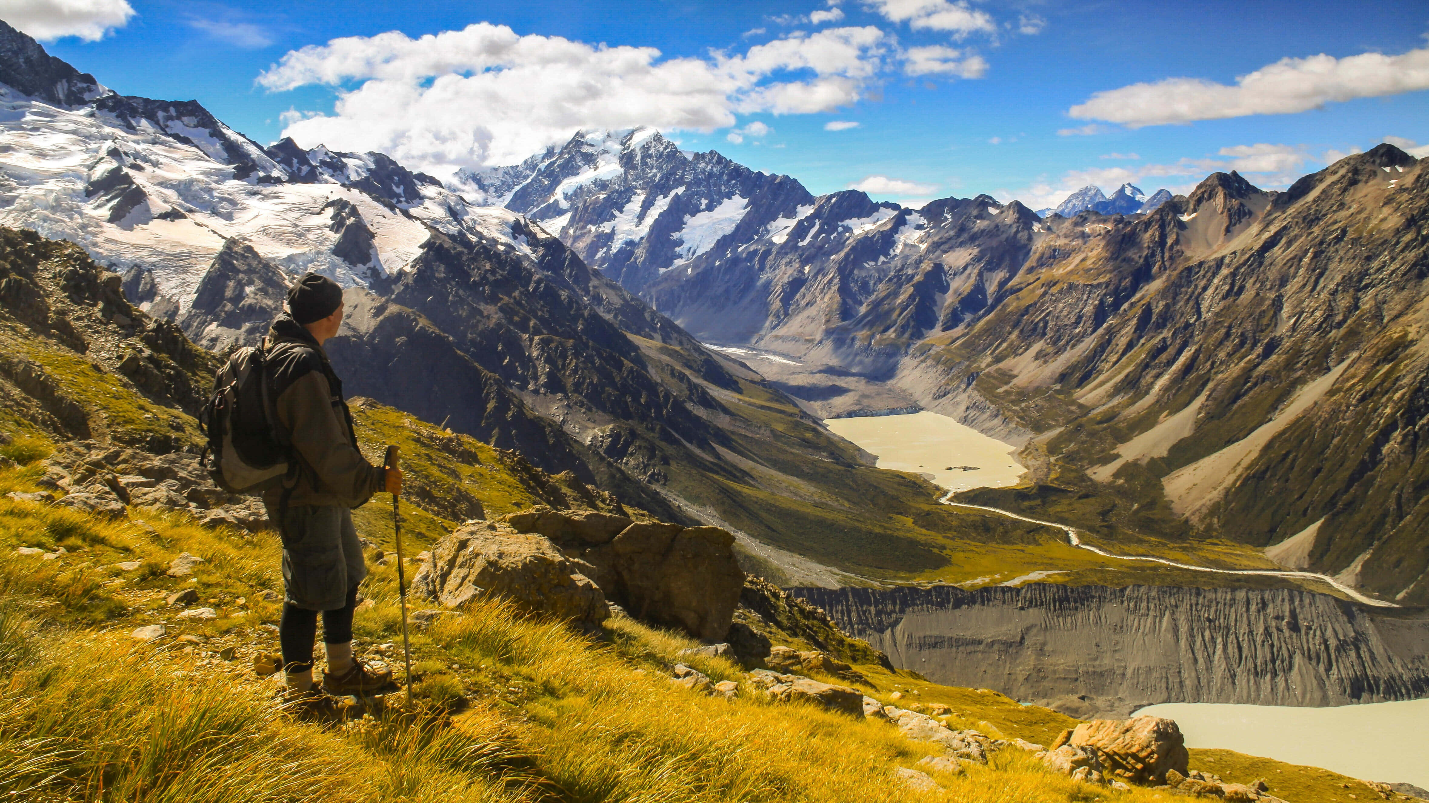 New Zealand 桌布 壁紙 — 一位揹著背包的遠足者站在長滿草的山脊上，俯瞰著一個山谷，山谷中有冰川湖和白雪覆蓋的山峰，天空湛藍明亮。
