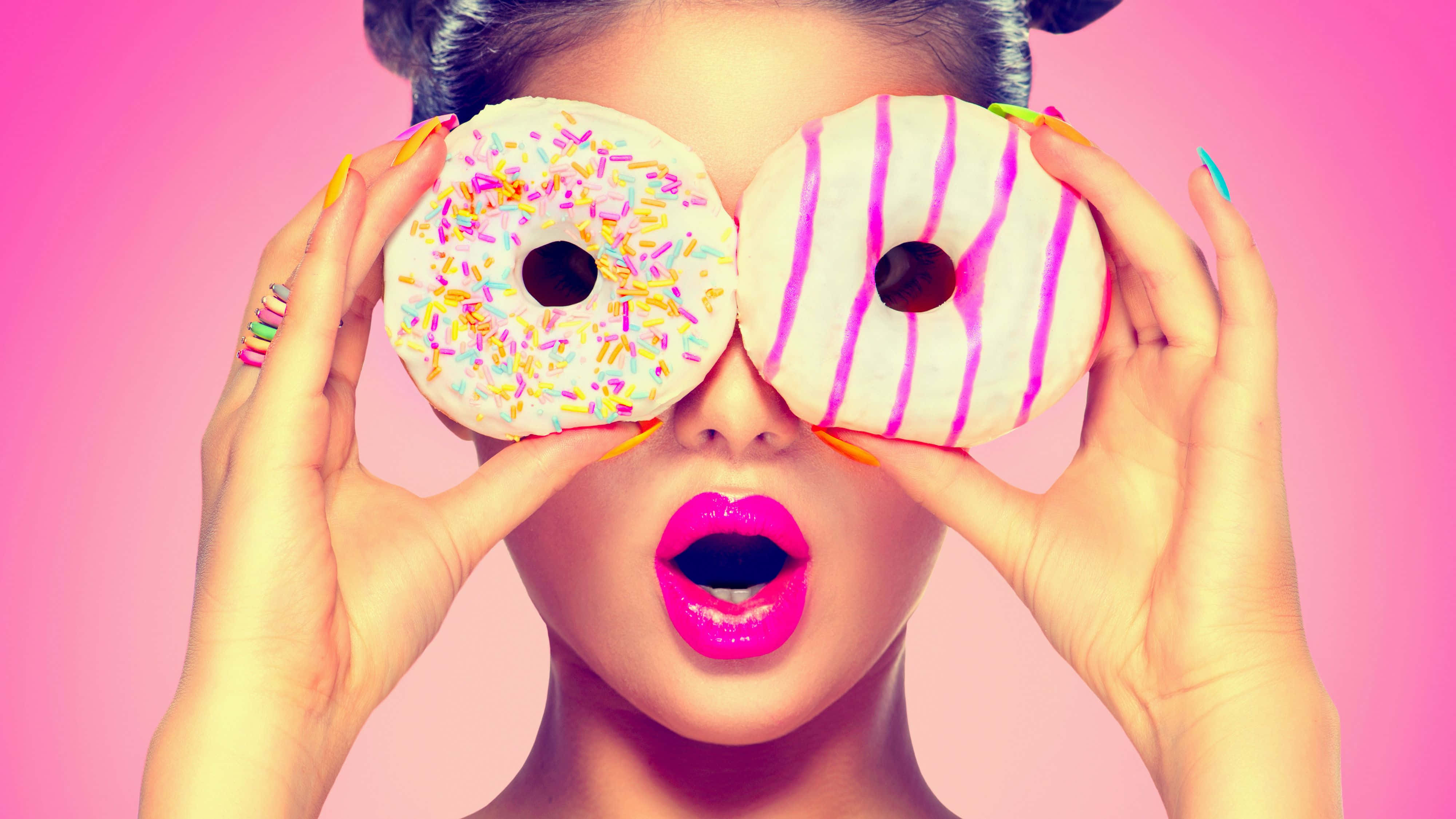 donuts 桌布 壁紙 — 一位擁有亮粉紅色嘴唇的女性，在鮮豔的粉紅色背景前，將兩個裝飾過的冬甩舉在眼前。