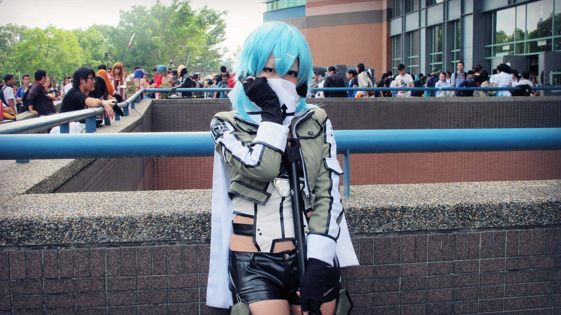 Sword Art Online Cosplay 桌布 壁紙 — 一位擁有亮藍色頭髮和白色面具的角色扮演者，在戶外展會上穿著精緻服裝擺姿勢，藍色欄杆後方有群眾。