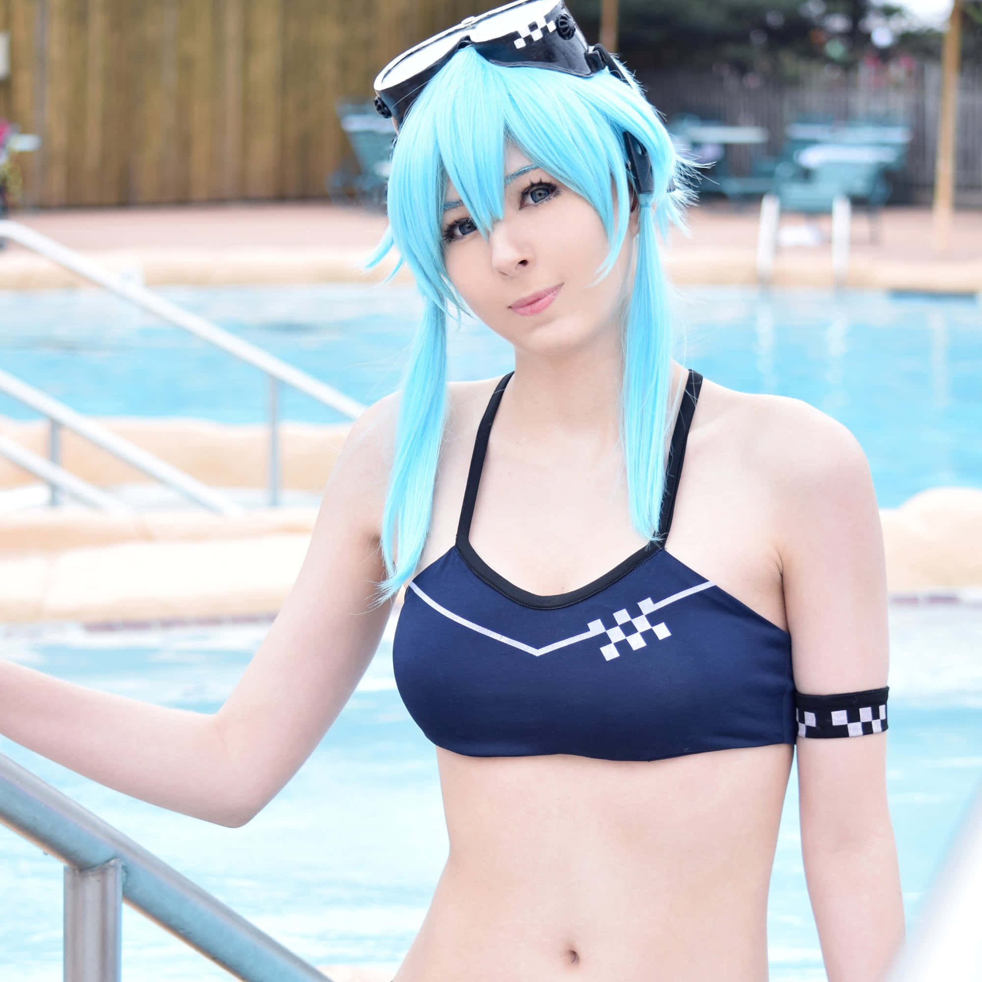 Sword Art Online Cosplay 桌布 壁紙 — 一位擁有亮藍色頭髮和賽車護目鏡的角色扮演者，身穿帶有白色格紋點綴的海軍藍泳衣，在泳池旁擺姿勢。