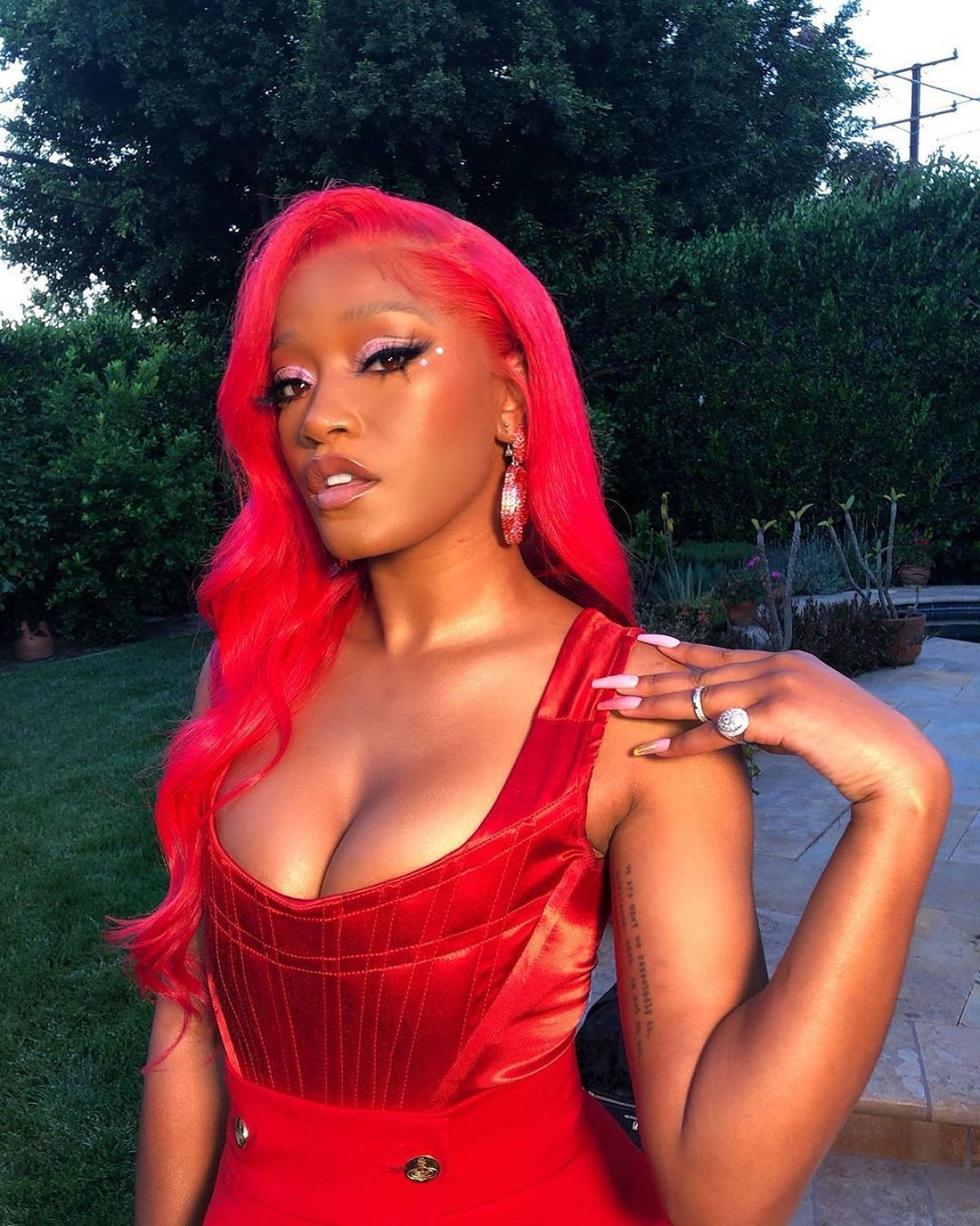 Keke Palmer《Sticky》 壁紙 — 一位擁有充滿活力的粉紅色頭髮和大膽妝容的女性，在鬱鬱蔥蔥的綠色花園背景下自信地擺出紅色連衣裙的姿勢。