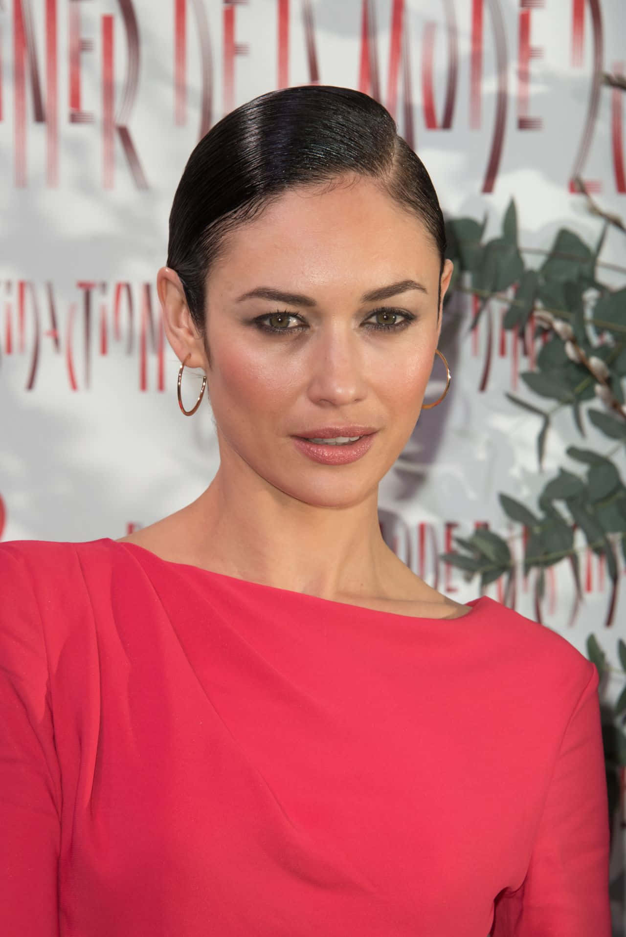 Olga Kurylenko 桌布 壁紙 — 一位擁有光滑黑髮和耳環的女性，在紅地毯活動中穿著鮮豔的粉紅色連衣裙，背景有綠植和文字。
