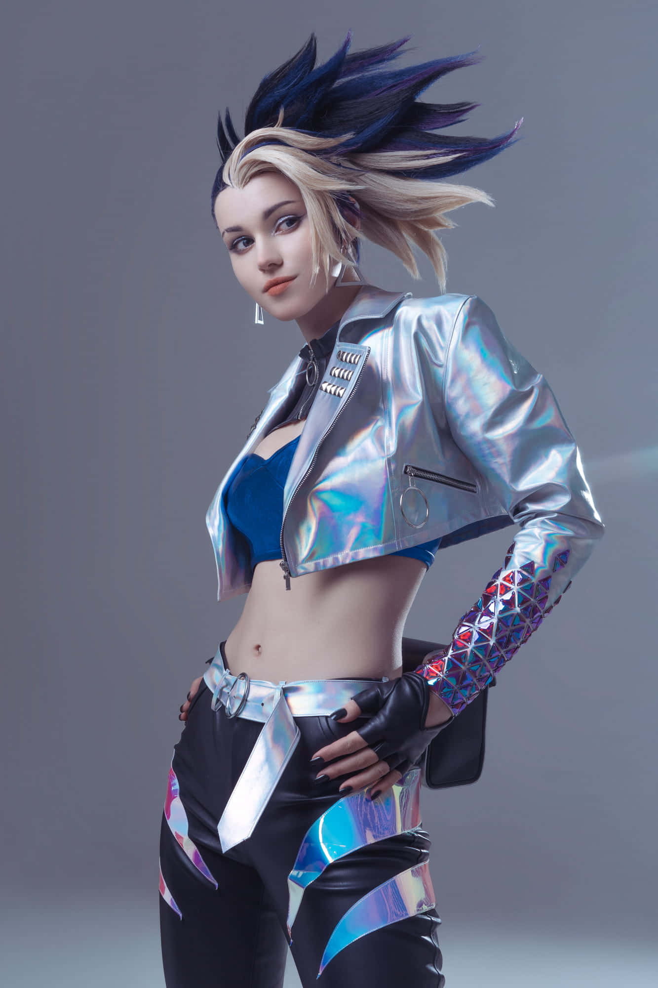 Gaming Cosplay 桌布 壁紙 — 一位擁有尖刺藍色和金色頭髮的角色扮演者，穿著閃亮的銀色外套和帶有全息圖案的黑色褲子，在灰色工作室背景前自信地擺姿勢。