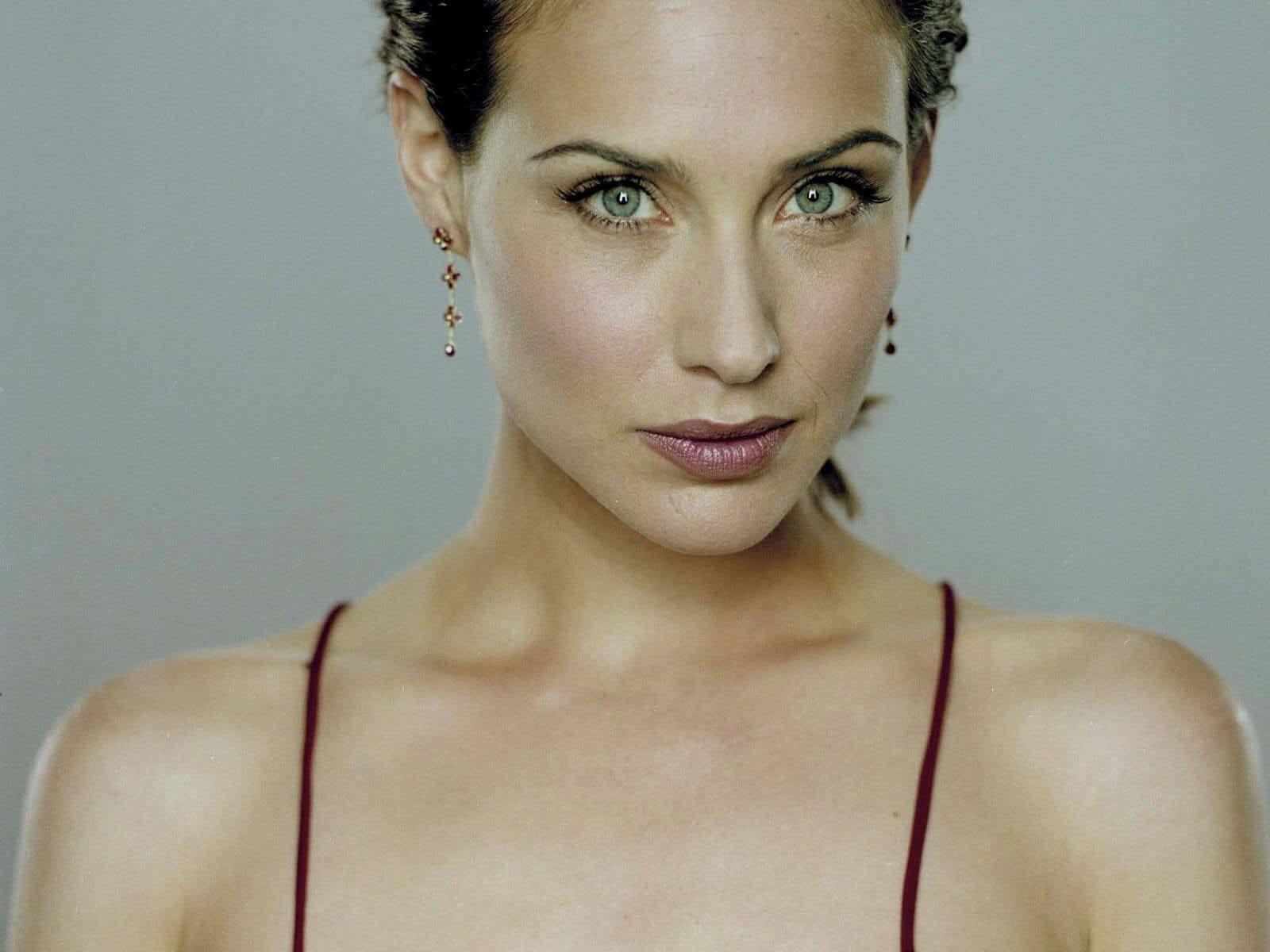 Claire Forlani 桌布 壁紙 — 一位擁有引人注目的綠色眼睛和向後梳的深色頭髮的女性特寫肖像，穿著紅色吊帶上衣和垂墜耳環，背景為中性灰色。