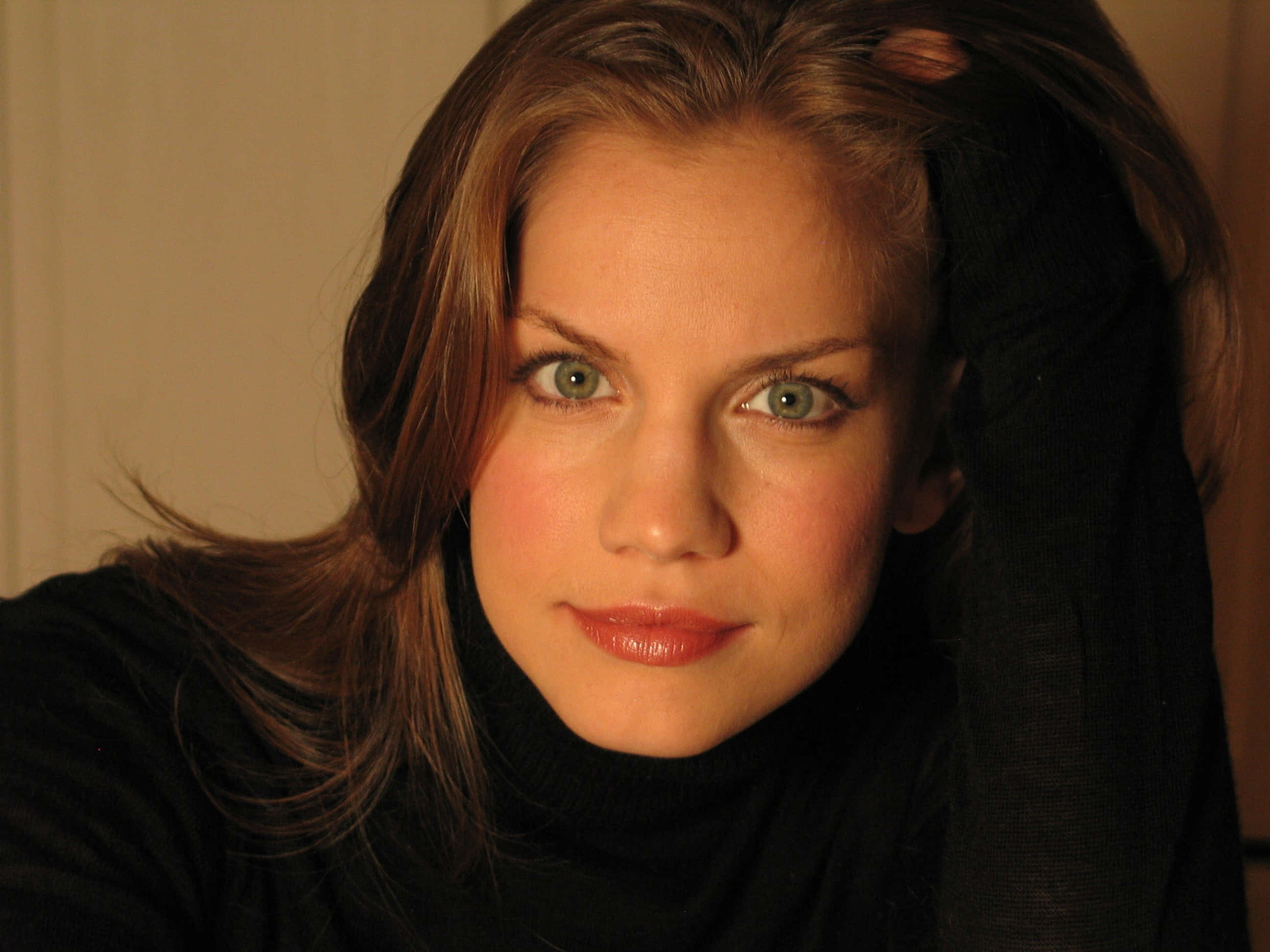 anna chlumsky 桌布 壁紙 — 一位擁有引人注目的綠色眼睛和棕色頭髮的女子的特寫肖像，穿著黑色高領毛衣，被溫暖的金色光線照亮。