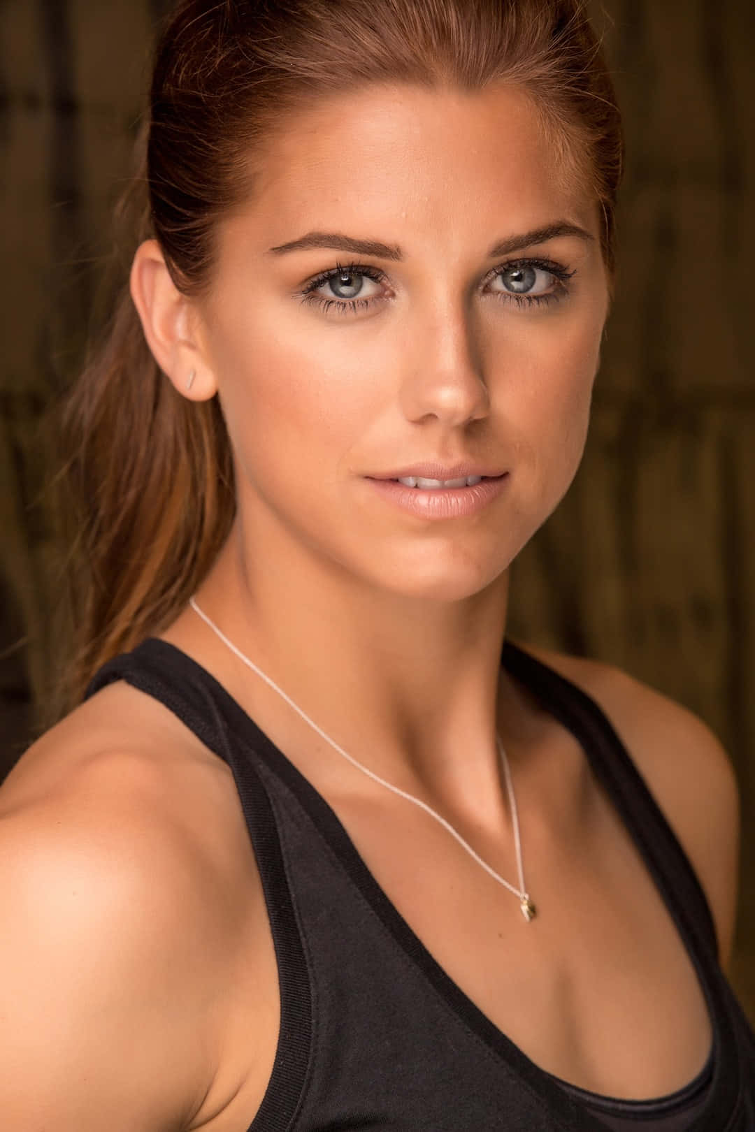 Alex Morgan 桌布 壁紙 — 一位擁有引人注目的藍色眼睛和棕色馬尾辮的女性特寫肖像，身穿黑色背心和精緻項鍊。