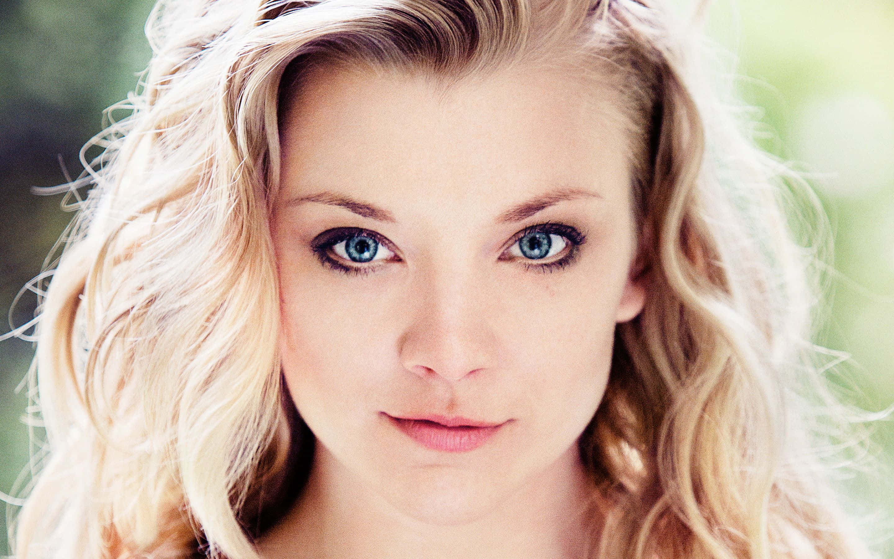 natalie dormer 桌布 壁紙 — 一位擁有引人注目的藍色眼睛和波浪金髮的女性，在柔和的綠色模糊背景下的特寫肖像。