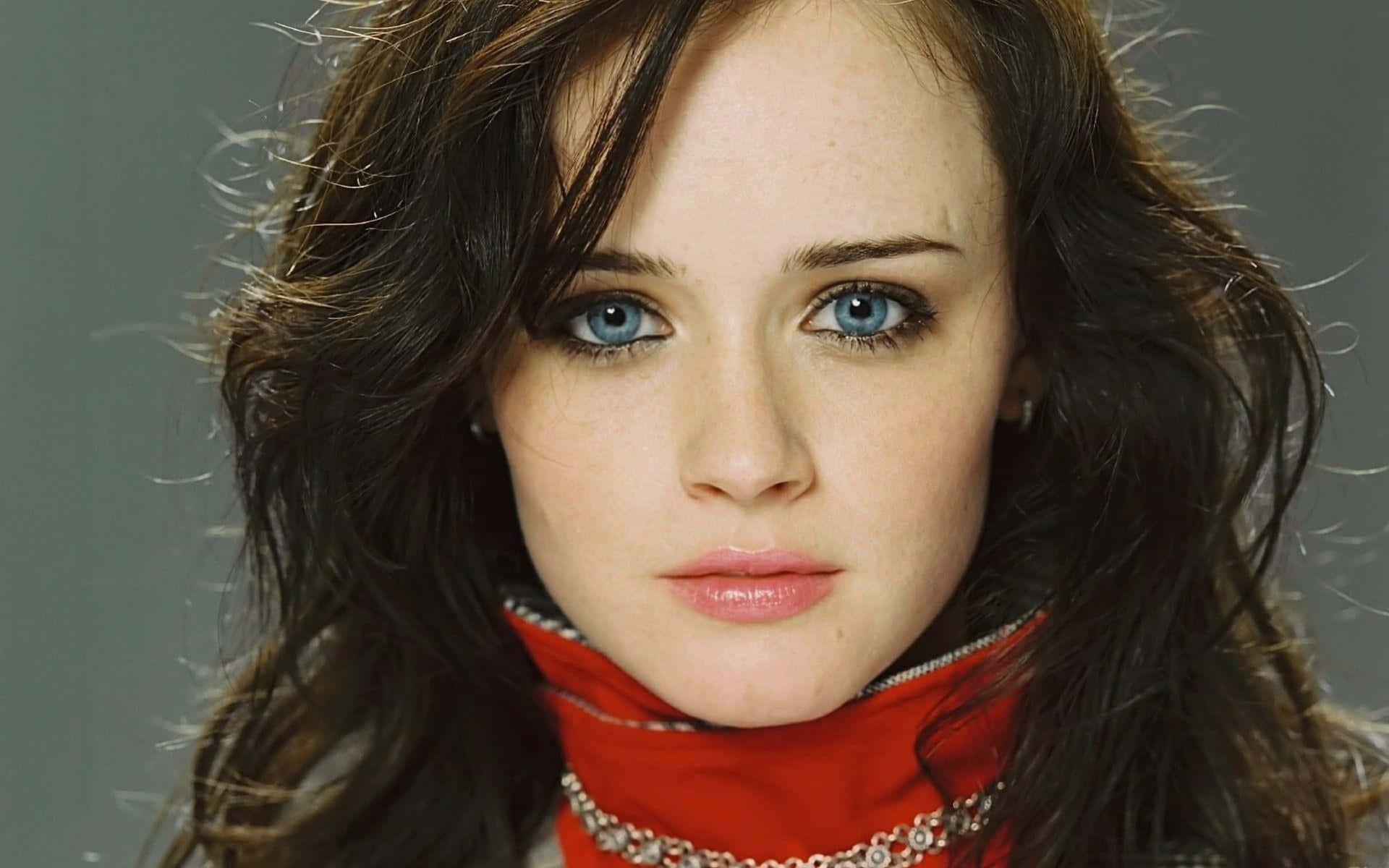 Alexis Bledel 桌布 壁紙 — 一位擁有引人注目的藍色眼睛和深色波浪髮的女性的特寫肖像，穿著帶有銀色裝飾的紅色高領毛衣。