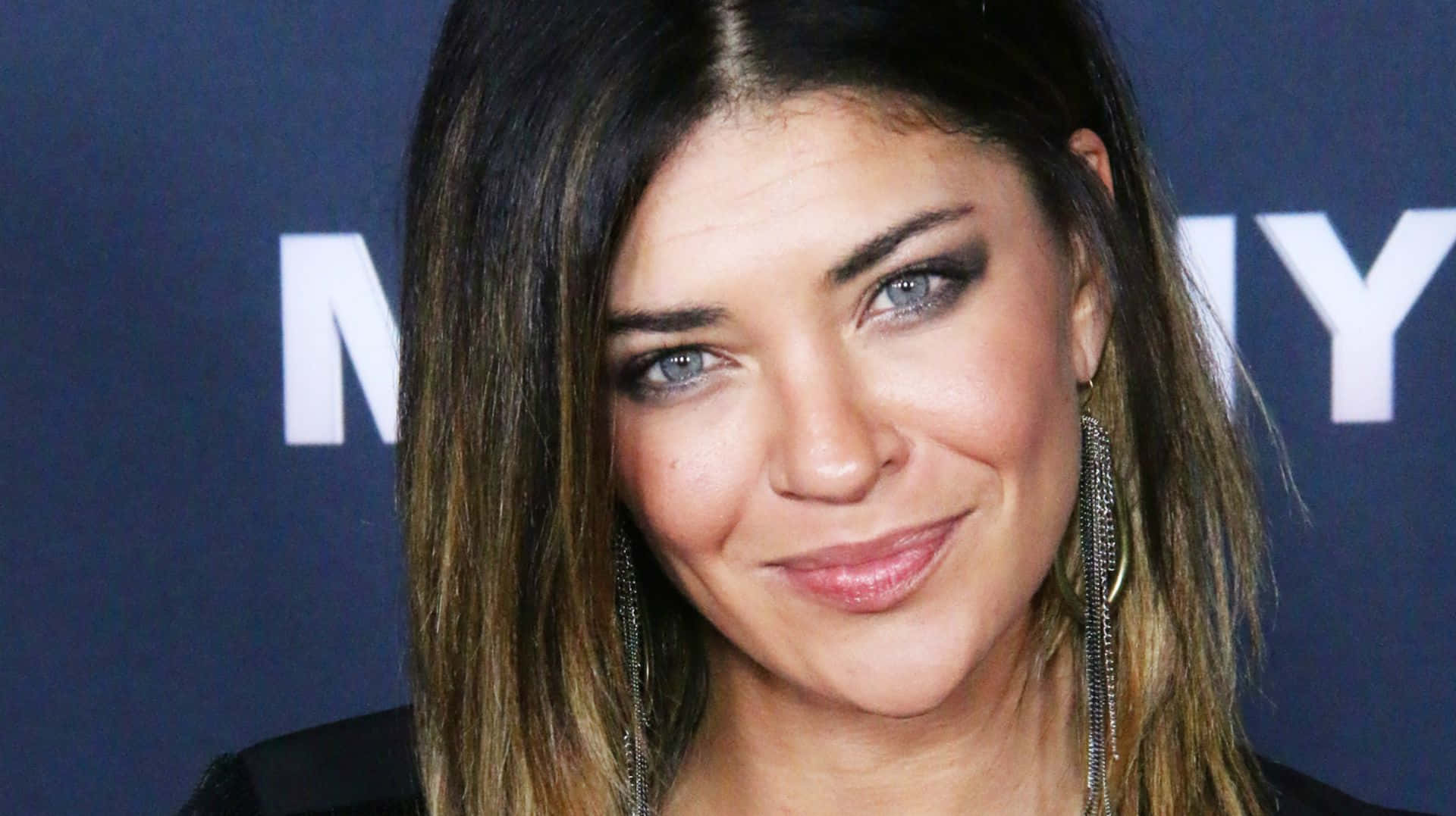 jessica szohr 桌布 壁紙 — 一位擁有引人注目的藍色眼睛和長層次頭髮的女性特寫肖像，在帶有白色文字的深藍色背景前溫暖微笑。