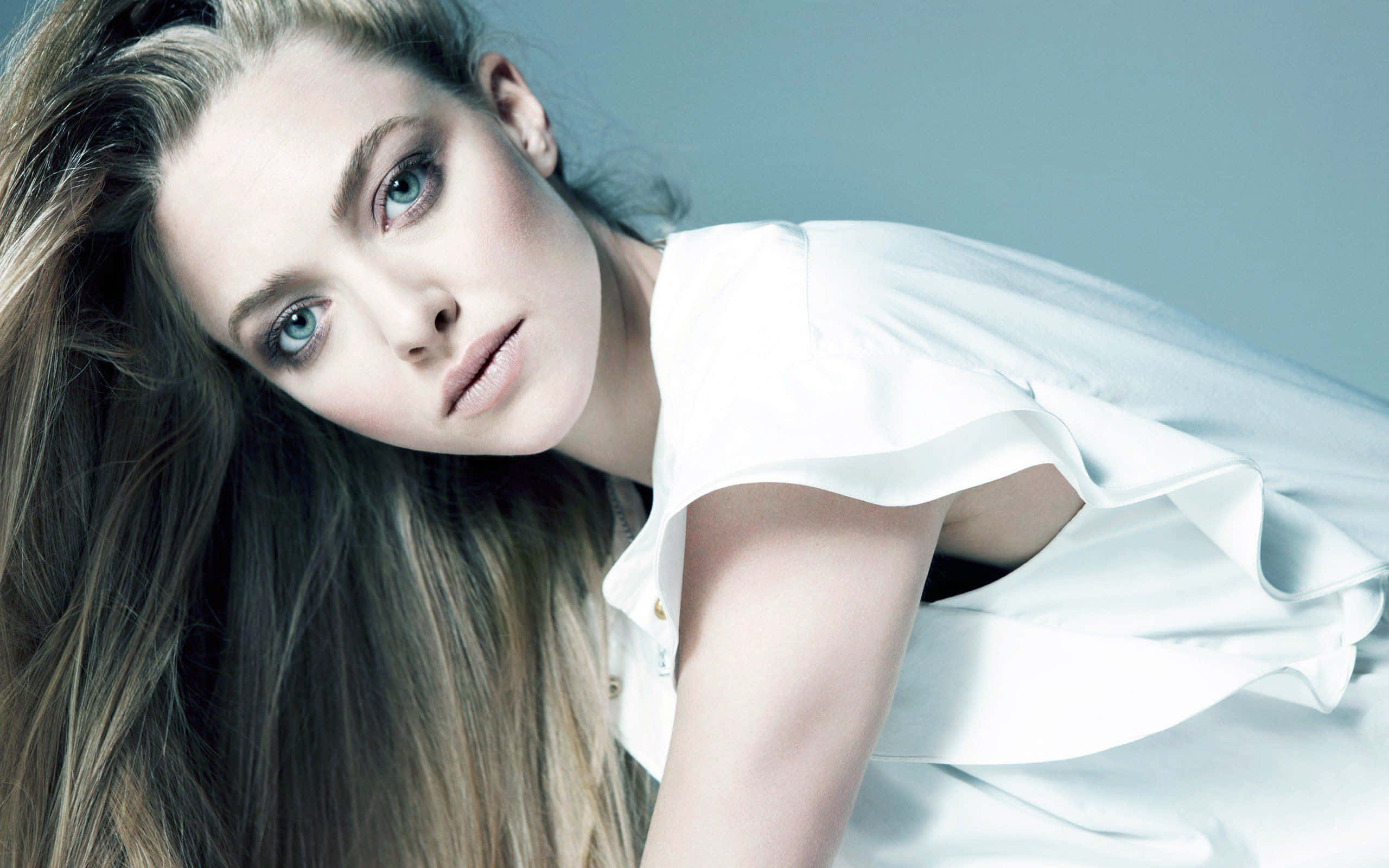 Amanda Seyfried 桌布 壁紙 — 一位擁有引人注目的藍色眼睛和飄逸金髮的女性，穿著白色襯衫，在柔和的藍色背景下凝視著。