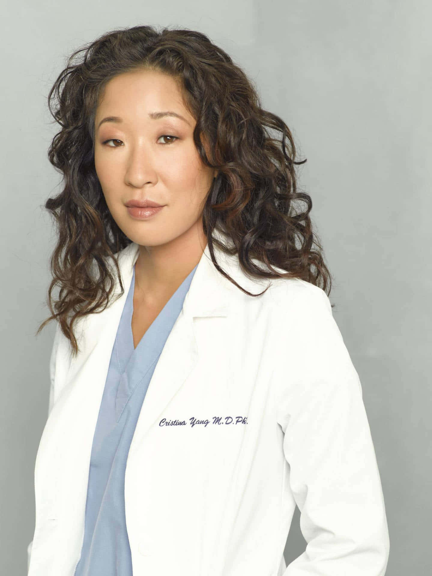 Grey's Anatomy 桌布 壁紙 — 一位擁有捲曲深色頭髮的女性，在純灰色背景前，穿著藍色刷手服，外罩白色實驗室外套。