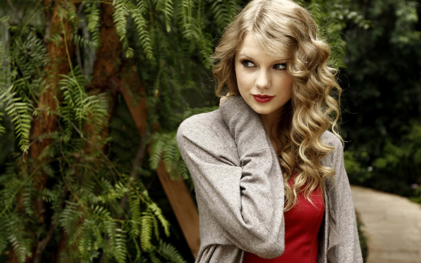 Taylor Swift 桌布 壁紙 — 一位擁有捲曲金髮和紅唇的女性站在一個綠意盎然、長滿蕨類植物的花園中，身穿灰色開襟衫，內搭紅色上衣。