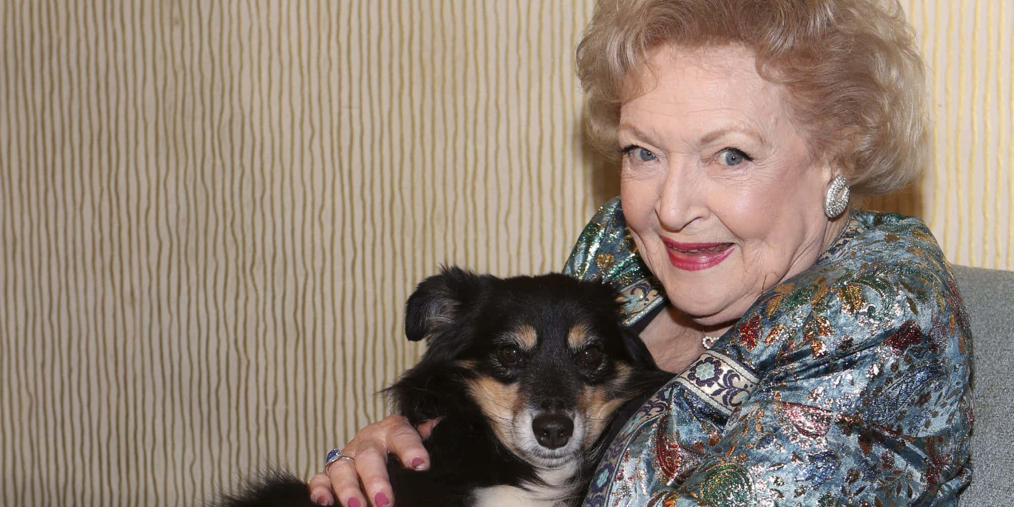 Betty White 桌布 壁紙 — 一位擁有捲曲金髮的老年女性，在條紋壁紙背景前，溫暖地微笑著擁抱一隻小黑褐色狗。