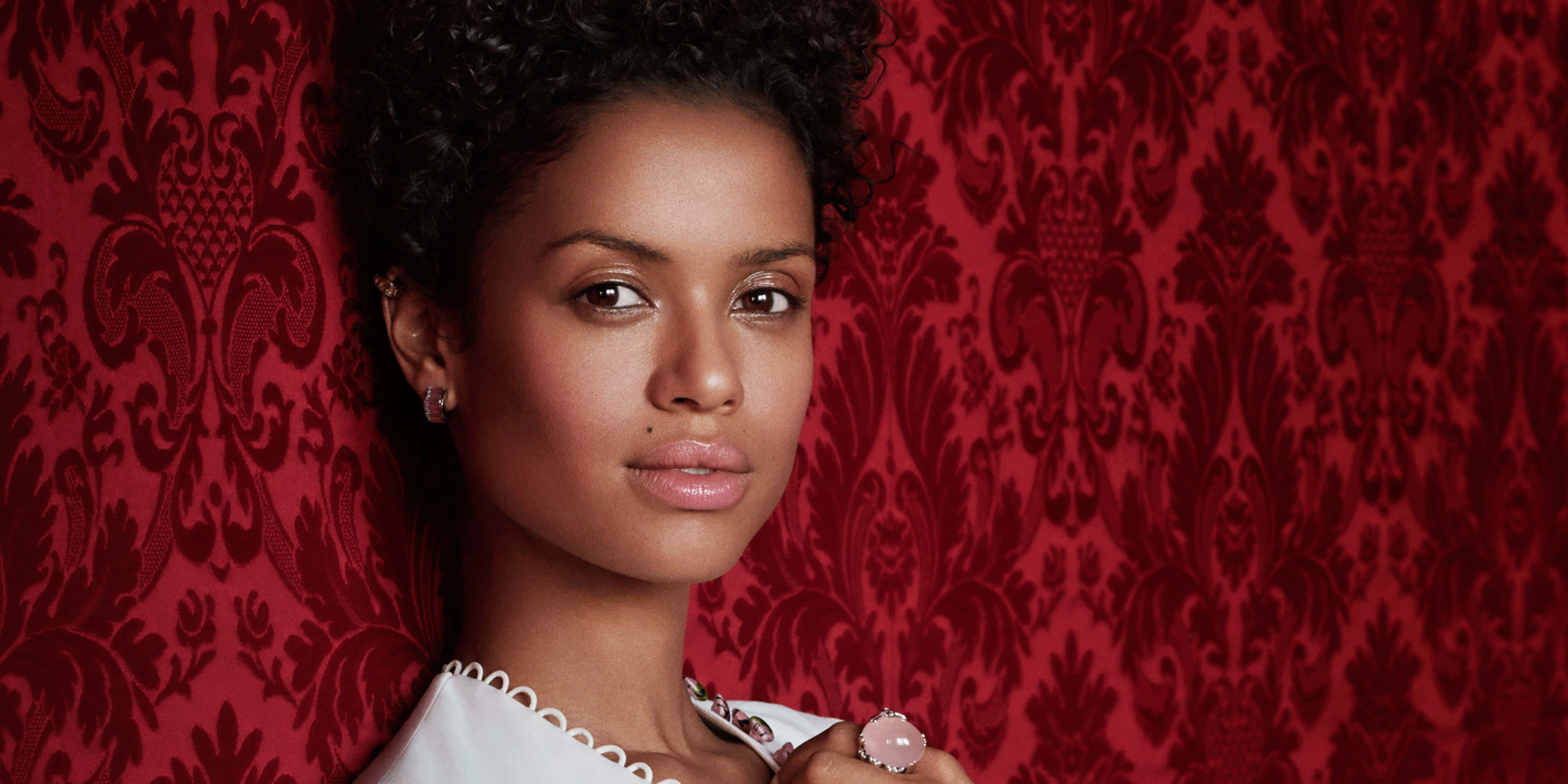 Gugu Mbatha-raw 桌布 壁紙 — 一位擁有捲髮的優雅女性，在華麗的紅色錦緞壁紙背景下溫柔凝視。