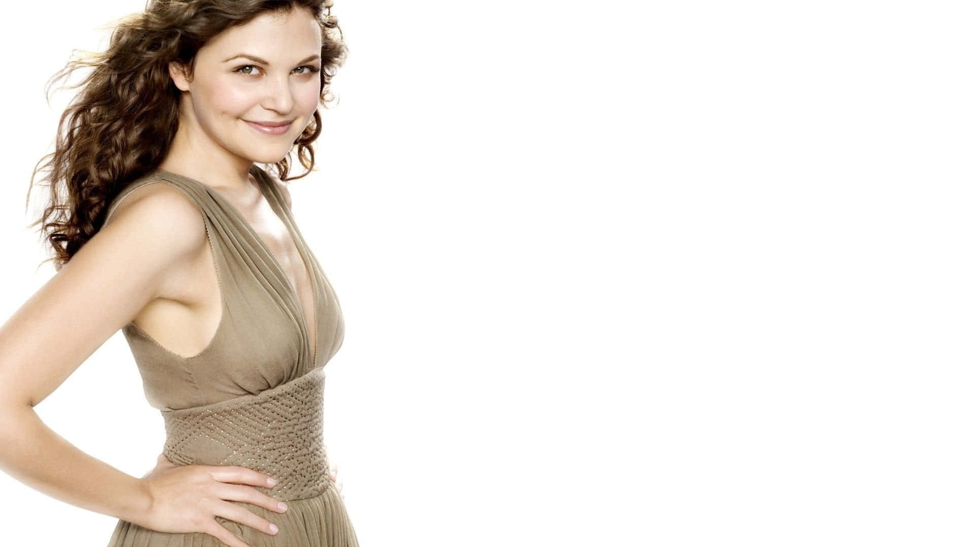 ginnifer goodwin 桌布 壁紙 — 一位擁有棕色捲髮的女性，身穿灰褐色連衣裙，在純白背景前自信微笑。